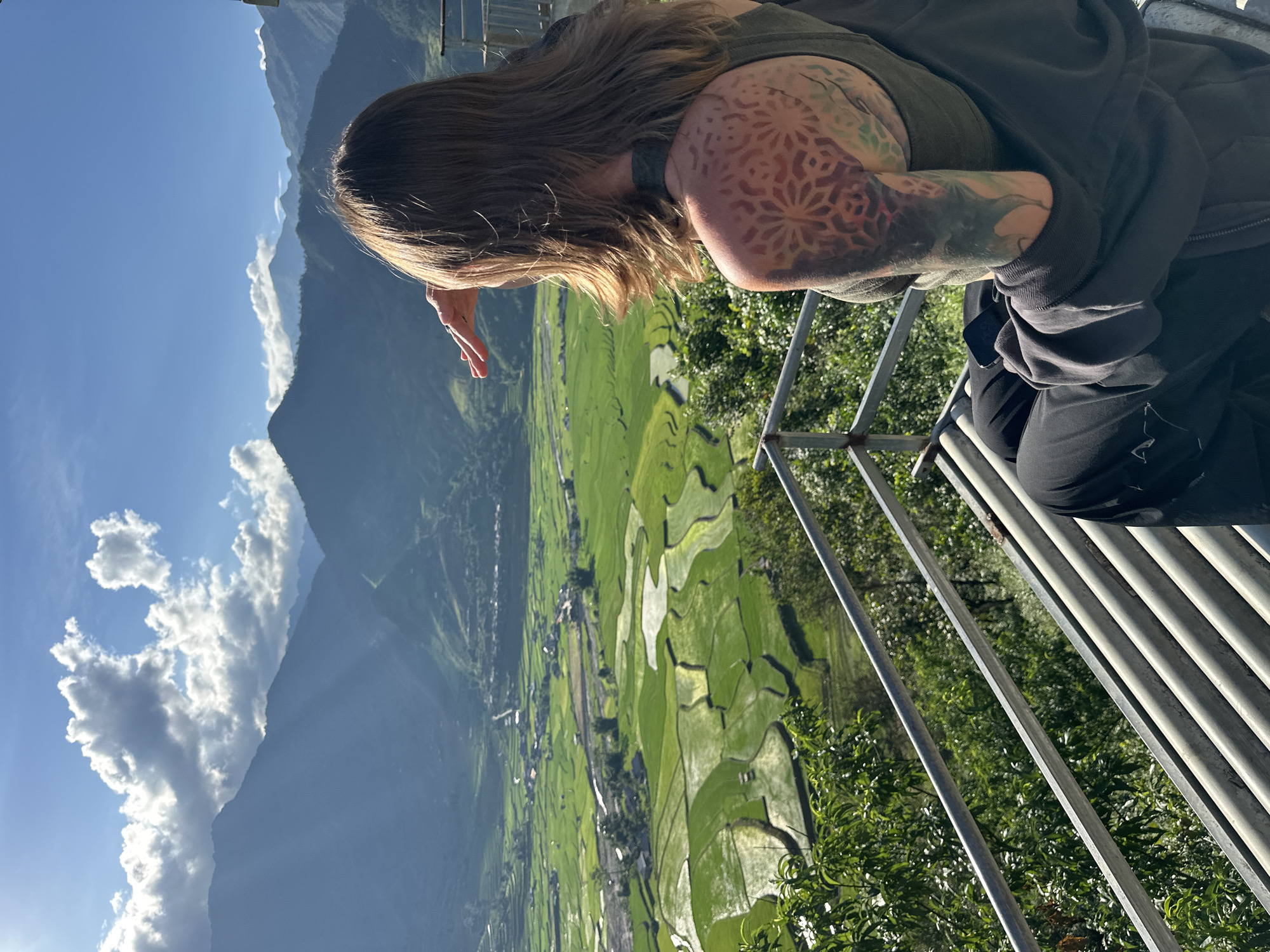 A person, seen from behind and the right side, is seated on a metal slatted bench on a viewing platform, observing a valley in Tú Lệ, Vietnam. The individual has long brown hair and prominent floral/geometric tattoos in red, orange, and green on their right shoulder blade and upper arm. They are wearing a dark grey tank top and a dark hooded jacket over black pants, with a mobile phone resting on their lap. Their right hand is raised, shielding their eyes from the bright sunlight. The panoramic view showcases a wide valley filled with vibrant green, irregularly shaped, and partially submerged rice paddies, indicating terraced cultivation. A winding river flows through the valley, dotted with small settlements and roads. In the background, multiple layers of dark mountains rise under a bright blue sky with scattered white cumulus clouds, from which visible sunbeams descend onto the mountain slopes. A metal railing is visible in the lower right and foreground of the platform.