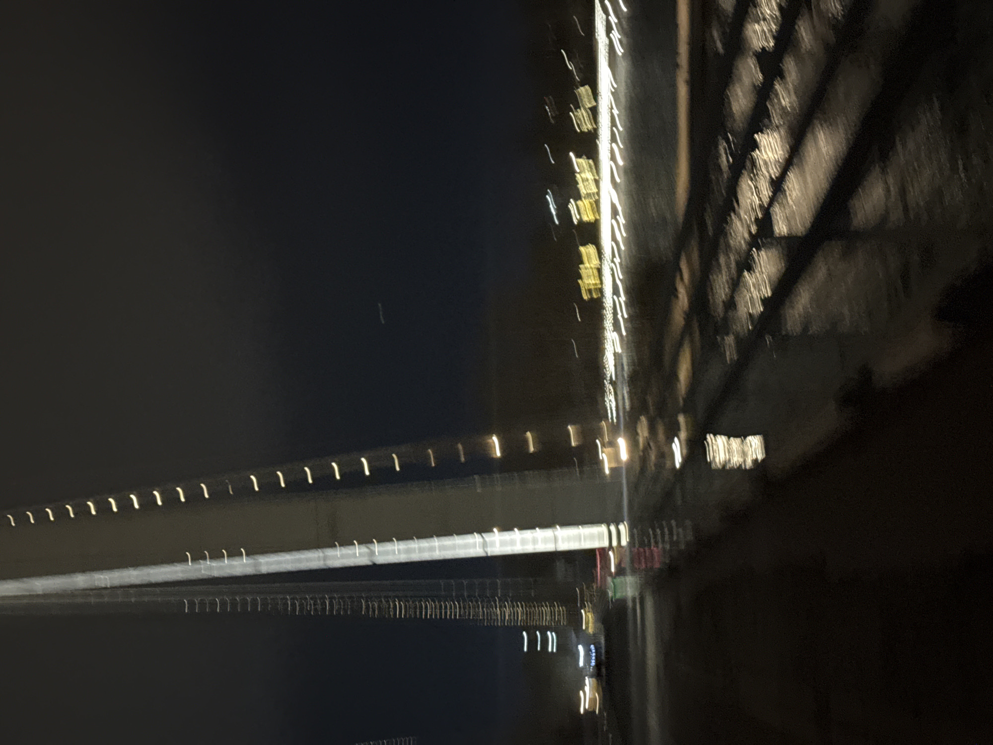 The image shows a night scene in Krasnoyarsk, Russia, likely taken with a long exposure and some motion. It captures the blurred lights of a bridge structure spanning a dark body of water. The lights create horizontal streaks, suggesting movement. The bridge has regular, repeating lights along its edge. There are shadowy structures on the right side of the frame, possibly related to the bridge construction or nearby buildings. The overall ambiance is dark and urban.