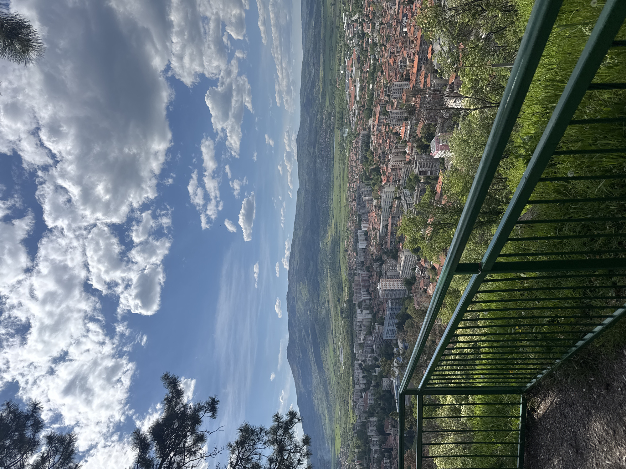 An elevated, eye-level view overlooks the city of Kyustendil, Bulgaria, under a sky filled with white clouds and blue sky. The perspective is from behind a green metal guardrail, positioned on a grassy area. The city, with its buildings and infrastructure, is spread out across the landscape, nestled against a backdrop of green fields and a mountain range. The foreground includes a portion of a tree with dark green foliage.