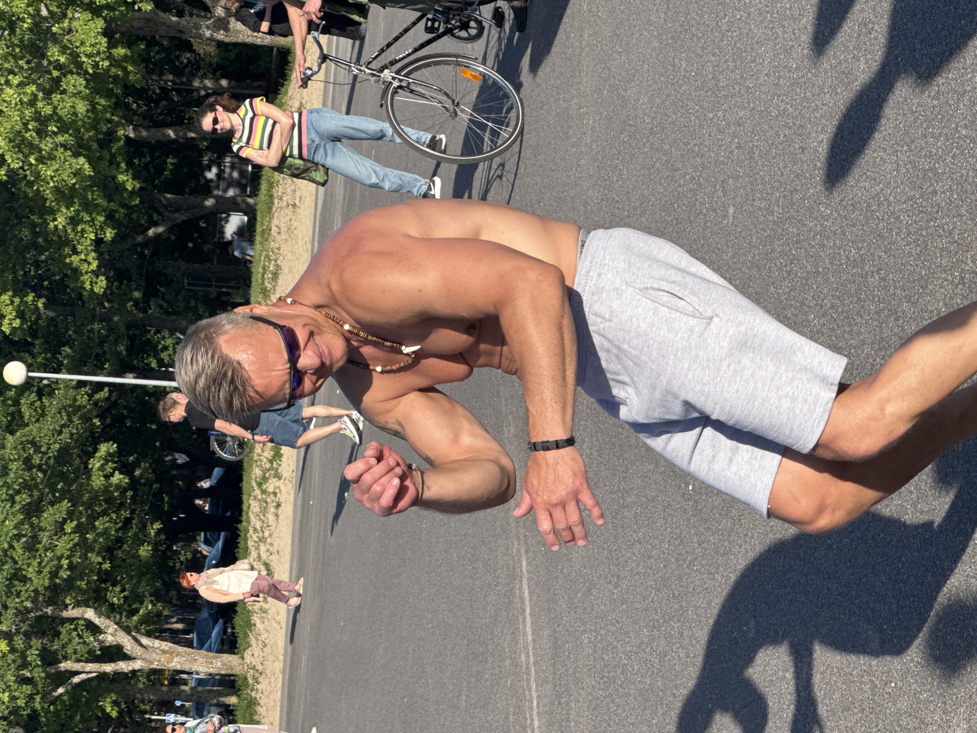 The image shows a shirtless man with gray hair and sunglasses dancing in a public outdoor space, possibly a park or a street in Tallinn, Estonia. He is wearing gray sweat shorts and a beaded necklace. In the background, other people are visible, some walking and some standing near a bicycle. The environment appears to be a paved area with trees and a sandy strip of land bordering it. The lighting suggests it is daytime, likely with a bright sun given the strong shadows. The man's pose and expression indicate he is actively engaged in dancing.