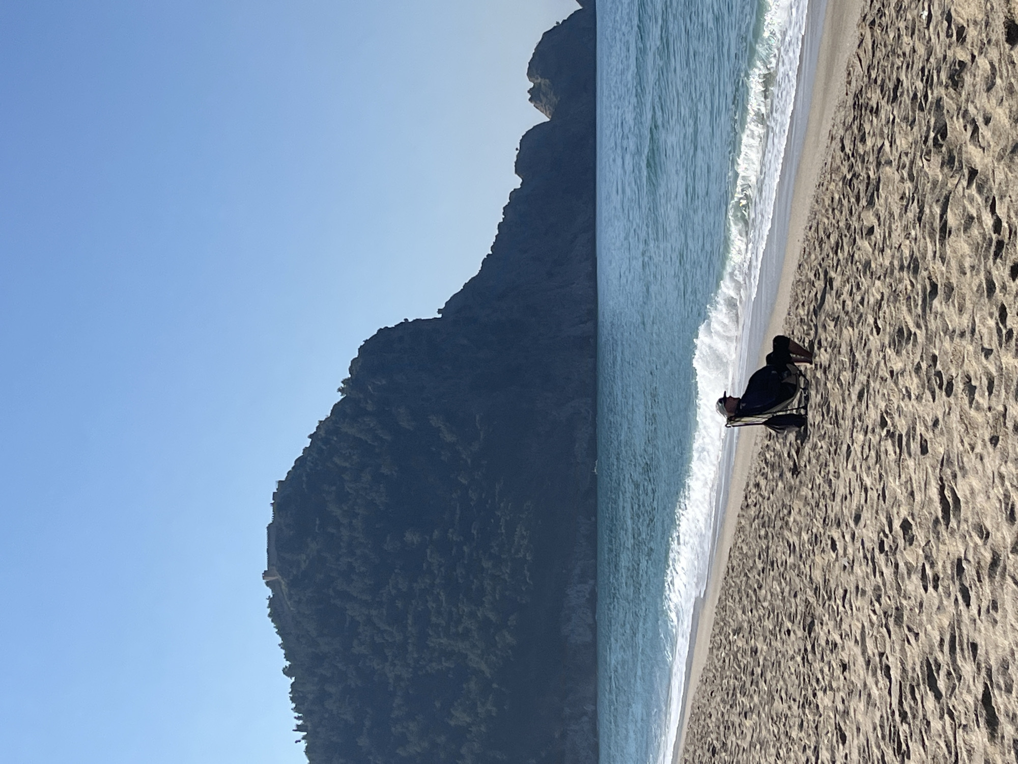 The image shows a beach scene in Alanya, Turkey, with a man sitting in a chair near the shoreline. The beach has light-colored sand and is bordered by a mountain covered in trees. The waves are white-capped and blue. The sky is clear and blue. The man is wearing dark clothing and appears to be relaxing.