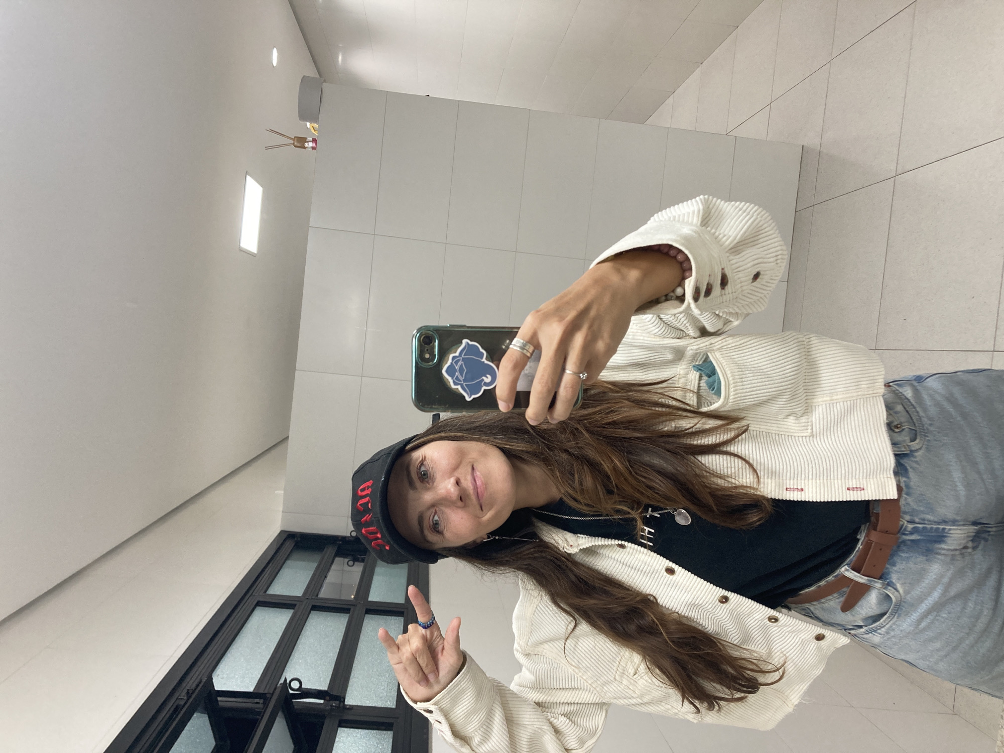 A woman is taking a selfie in a bathroom. She is wearing a black baseball cap with red lettering, a white corduroy jacket, a black t-shirt, and blue jeans. She has brown hair and is wearing several rings. She is holding her phone in her right hand and making a hand gesture with her left hand. The bathroom has white tiled walls and a black-framed window. The location is Florianópolis, Brazil.