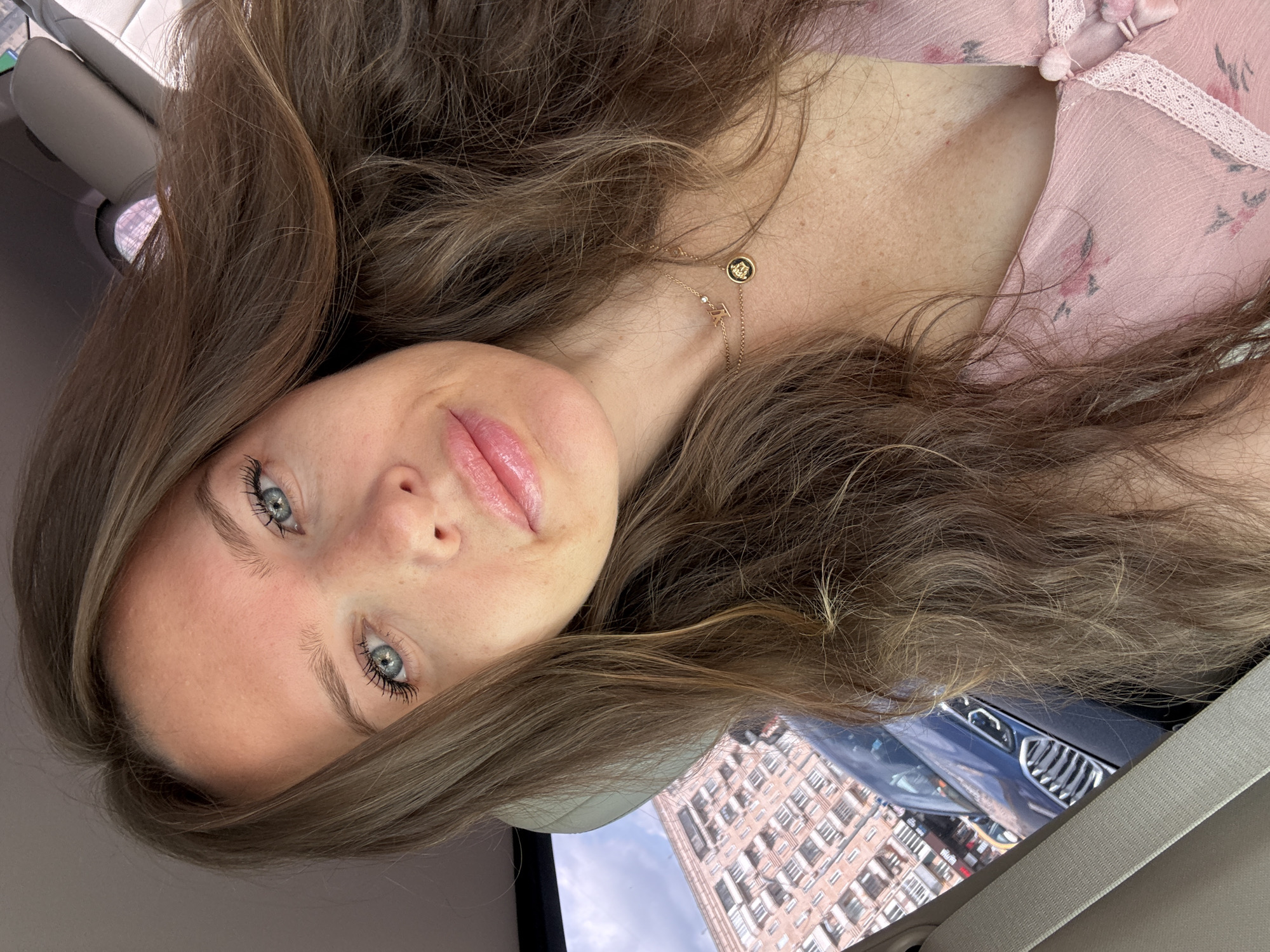 A medium shot captures a young woman with long, wavy brown hair inside a car. Her hair is styled loosely and falls around her face and shoulders. She has light blue eyes, fair skin with freckles, and plump, glossy pink lips. She is wearing a light pink dress with a floral pattern and a delicate lace trim. A gold necklace with a pendant is visible around her neck. Through the car window, a multi-story brick building with numerous windows and balconies is visible, suggesting an urban setting. The car's interior, including a gray seatbelt and headrest, is also in view. The overall impression is a selfie taken in an urban environment.