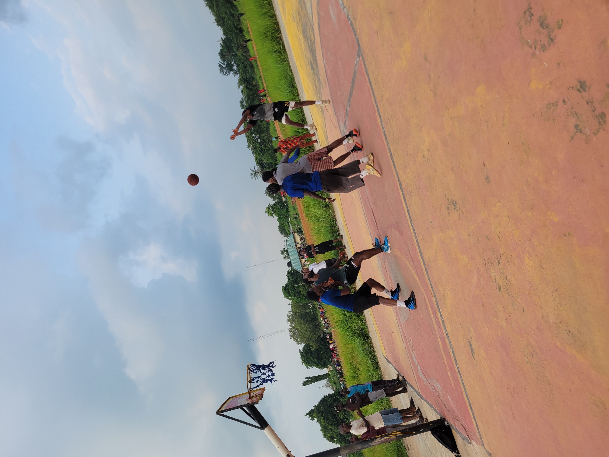 An outdoor basketball game is in progress on a reddish-orange and yellow-painted concrete court in Egbagu, Nigeria, under an overcast sky. The court surface shows signs of wear, including dark stains and faded paint lines. A brown basketball is captured in mid-air above the right side of the court. A player wearing a black and white patterned jersey with the number "24" on black shorts is airborne, having just released the ball towards an unseen basket. He is wearing white athletic shoes.
Three other players are in proximity to the airborne shooter. An opposing player wearing a blue t-shirt, black shorts, and blue and black athletic shoes is positioned directly in front of the shooter. To the left of this defensive player stands another player in a grey t-shirt, plaid shorts, and orange and black athletic shoes. Further towards the basket, a fourth player in a blue t-shirt, grey shorts, and light yellow athletic shoes is also on the court.
The basketball hoop, consisting of a white backboard, a black support pole, and a blue net, is visible on the left side of the court. A black backpack rests on the ground at the base of the basketball pole. Several individuals are observing the game from the sidelines; four men are standing near the basketball pole on the left, and a larger crowd is visible in the distant background. The court is surrounded by vibrant green grass and numerous trees. Distant buildings and utility poles are discernible beyond the treeline.