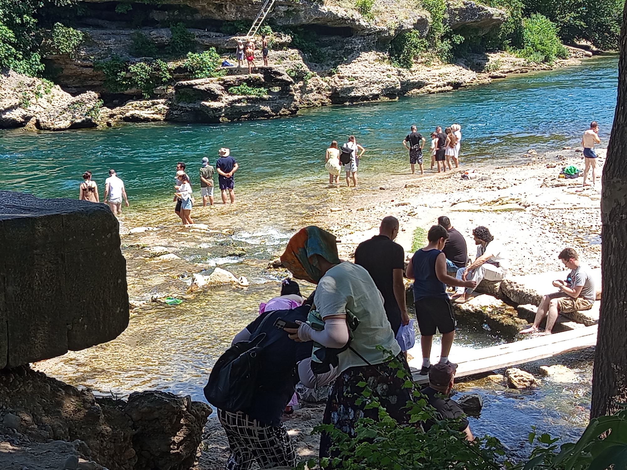 The image depicts a scenic view of the Bližanjka River in Mostar, Bosnia and Herzegovina, a popular natural attraction known for its clear turquoise waters and rocky surroundings. A group of people are engaged in various activities along the riverbank. Some are wading in the shallow water, while others are standing on the rocky shore. In the foreground, several individuals are dressed casually, some with backpacks, suggesting a day trip or recreational outing. One person in the center is wearing a headscarf and holding a smartphone. Further back, a group of people are gathered near the water's edge, with some appearing to be preparing to enter the river. In the background, a steep, rocky cliff face is visible, covered in lush green vegetation, with a wooden ladder leading to an elevated platform where a few individuals are standing. The river flows gently, with sunlight illuminating the clear water and the pebbles on the riverbed. The overall atmosphere is one of leisure and enjoyment of the natural environment.