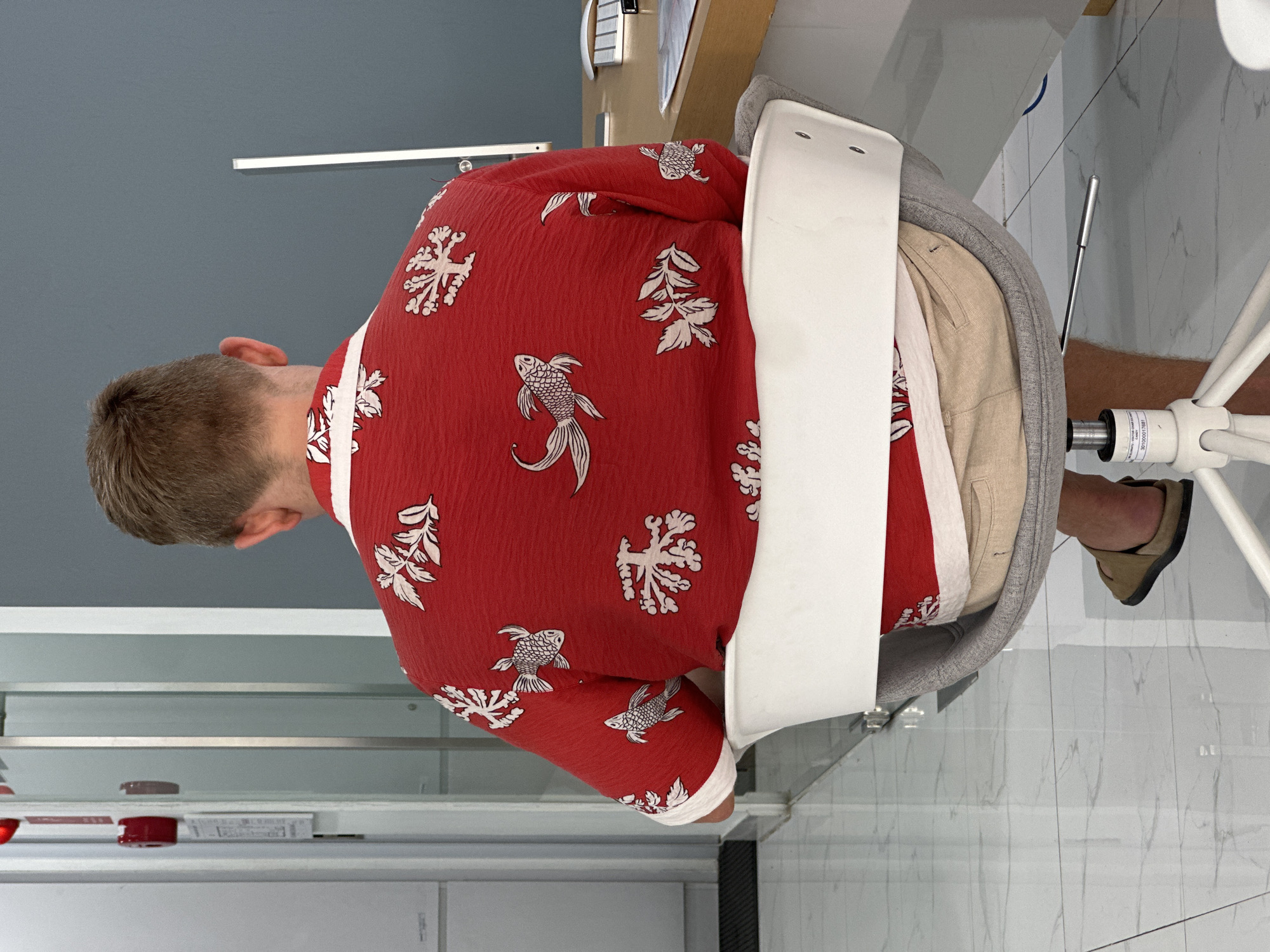 A man with short, light-brown hair is sitting in a grey office chair with a white back. He is wearing a red shirt patterned with white snowflakes and fish. He is also wearing light tan shorts and tan slip-on shoes. The environment includes a room with a light grey wall, a white cabinet with shelves, a desk with a beige top, and white marble-patterned floor. The scene appears to be indoors. The location is Kuta Selatan, Indonesia.