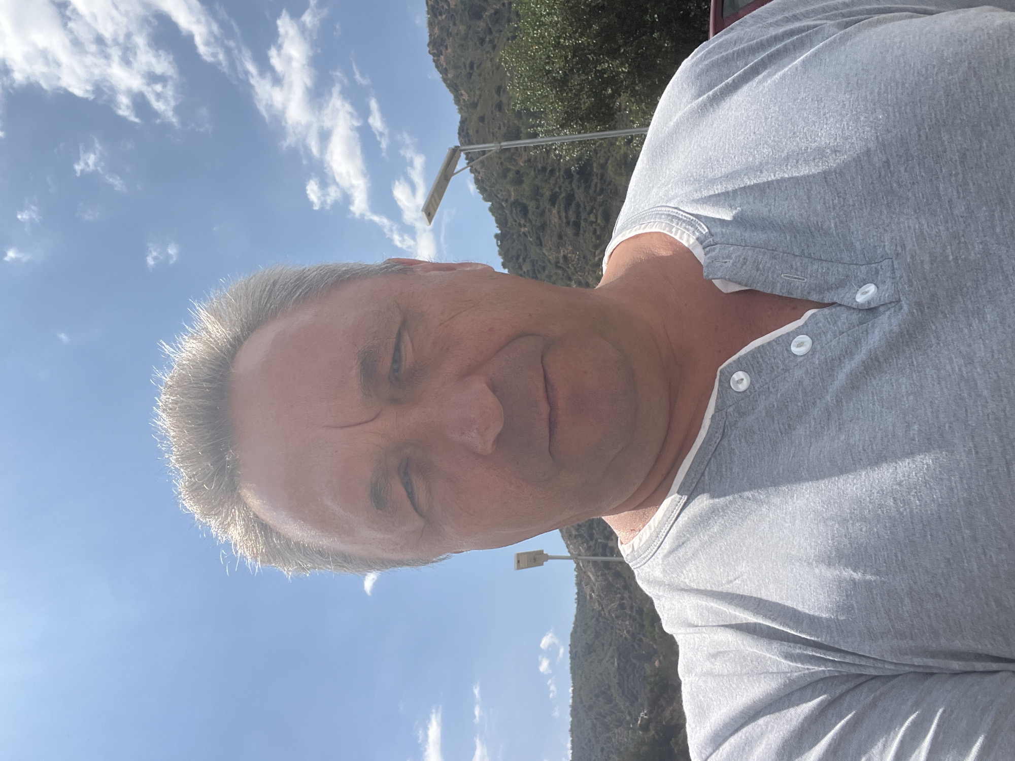 A medium-shot, selfie-style photo shows a middle-aged Caucasian male with light-colored hair and fair skin. He is wearing a light gray henley shirt with a white collar and two visible white buttons. His expression is neutral, with his eyes looking directly at the camera. The background features a bright blue sky with scattered white clouds. Behind him, a hilly, arid landscape is visible, dotted with sparse green vegetation. A modern solar-powered street light stands prominently on a pole to his right. The overall setting suggests an outdoor location, possibly in a rural or semi-urban area under bright sunlight. The location is identified as Kalopanagiotis, Cyprus.