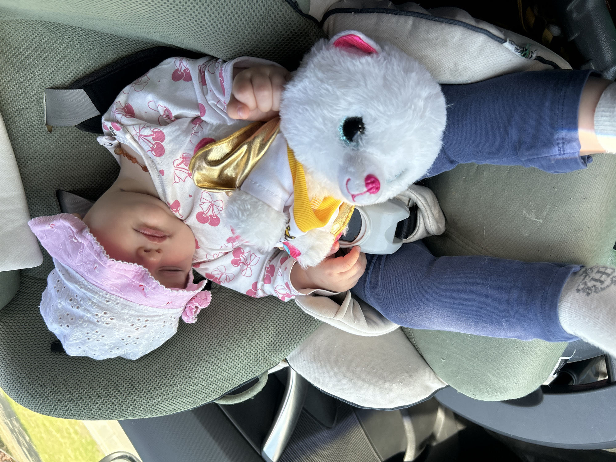 The image captures a baby sleeping peacefully in a car seat, accompanied by a white plush toy. The baby is dressed in a white, long-sleeved top with a pink cherry pattern, blue leggings, and white socks, and wears a light pink and white bonnet. A small, amber-colored beaded necklace is visible around the baby's neck.
The plush toy, which appears to be a white polar bear or cat, features sparkly blue eyes, pink ears, and a pink nose. It is adorned with a gold and yellow scarf or accessory and rests against the baby's body.
The baby is secured in an olive-green car seat with a mesh fabric back and lighter beige padded sections. Safety straps are visible across the baby's shoulders and chest. The setting is inside a car during daytime, with bright light illuminating the scene. Through the car window in the background, green grass or foliage is visible, suggesting the car is outdoors or in transit. A silver car door handle is partially seen. The overall atmosphere is calm and serene, with the baby appearing to be in a deep sleep. Faint text "jcb.com" is visible on a tag attached to the car seat padding. The image was taken in Moscow, Russia.