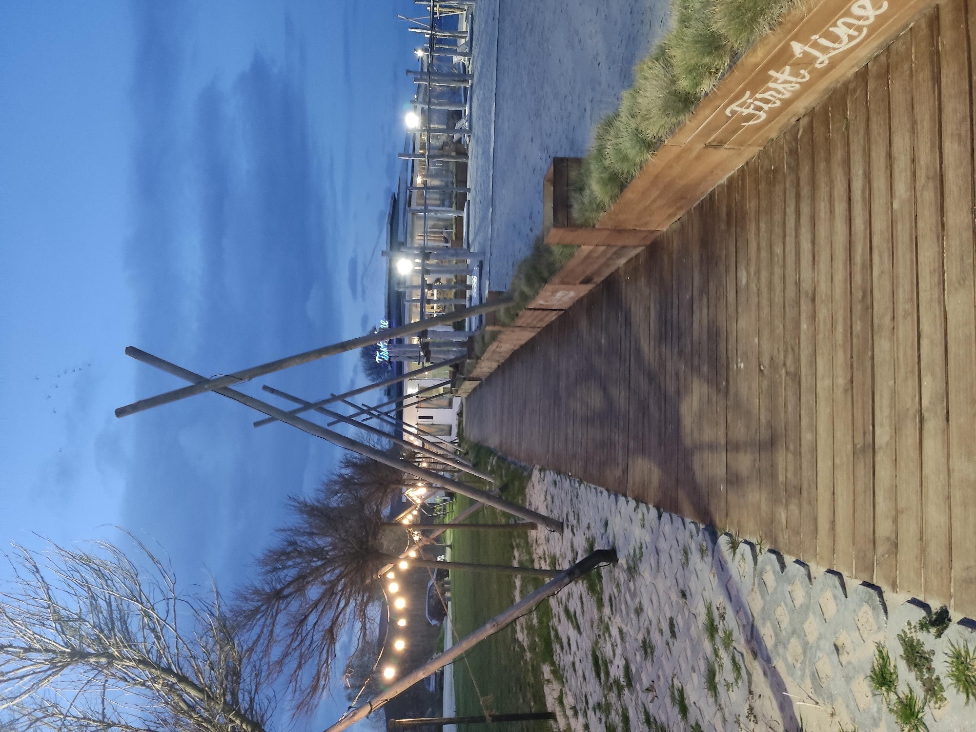The image captures an outdoor scene at dusk, likely in a public area, featuring a wooden boardwalk or pathway. The location includes a section of what appears to be a beachfront or grassy area. The phrase "First Line" is written in white on the wooden structure along the path.
Elements of the scene include:
- A wooden boardwalk or path.
- A grassy area with trees and what appears to be string lights hanging from the trees.
- A building or structure in the distance, with lights illuminated.
- The sky is dark blue, suggesting twilight.
- Some areas of sandy terrain are visible.
The specific location within the city of Unknown, Unknown, is not identifiable from this image alone. No people are visible in the photograph.