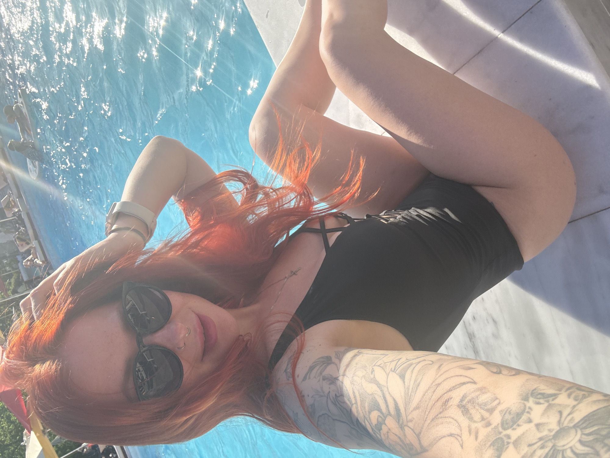 A woman lies on a white marble surface next to a pool of water, potentially in the city of Rostov-on-Don, Russia. The water reflects sunlight. She is wearing a black one-piece swimsuit and sunglasses. Her hair is red, and she has tattoos on her arm. She is holding her hair with her left hand. In the background, other people and structures are faintly visible.