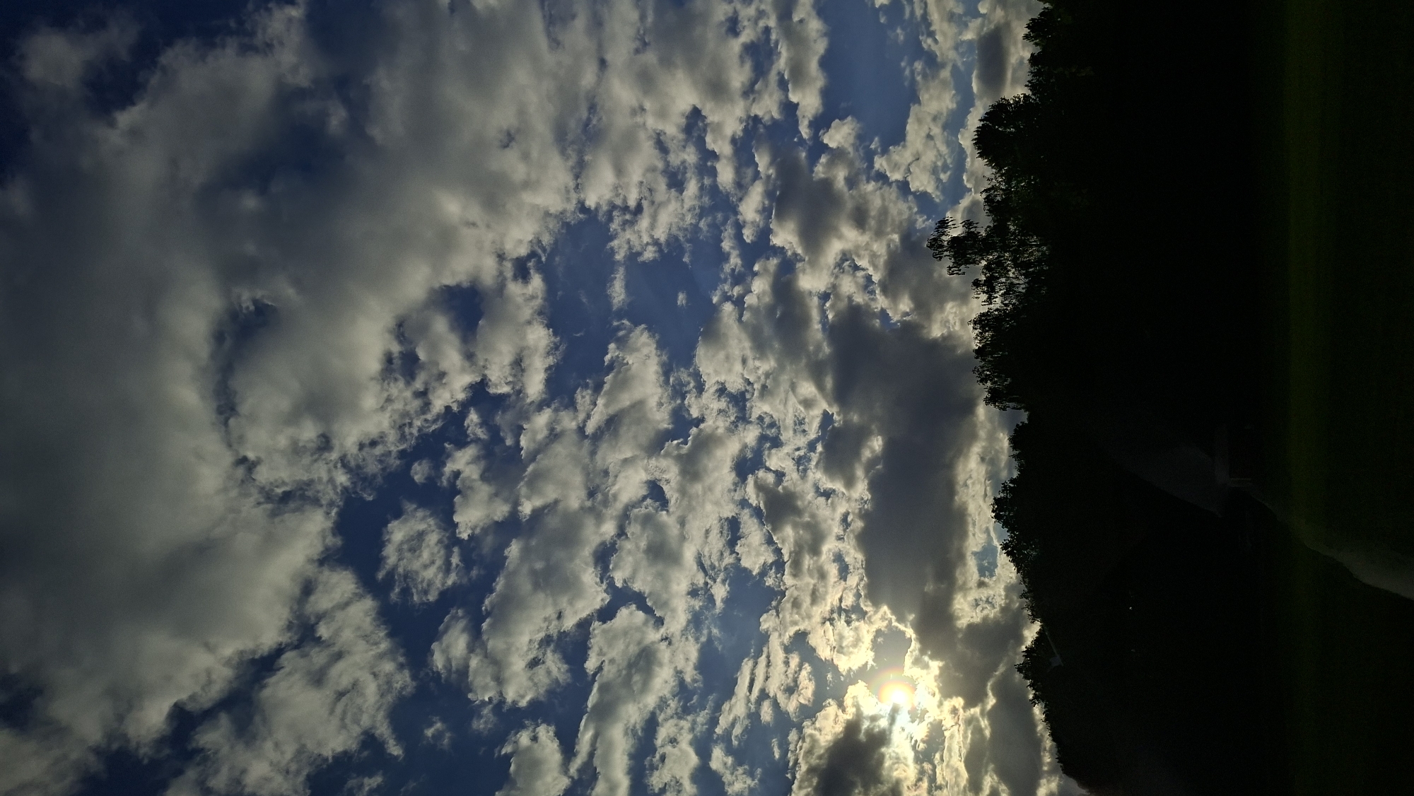The image shows a sky filled with scattered cumulus clouds, partially obscuring the sun, with the silhouette of trees along the right edge. The location is specified as Ilyanskiy selskiy Sovet, Belarus, but the image itself offers no visual confirmation of this location. There are no people or other obvious objects. The scene depicts a cloudy day with sunlight piercing through.