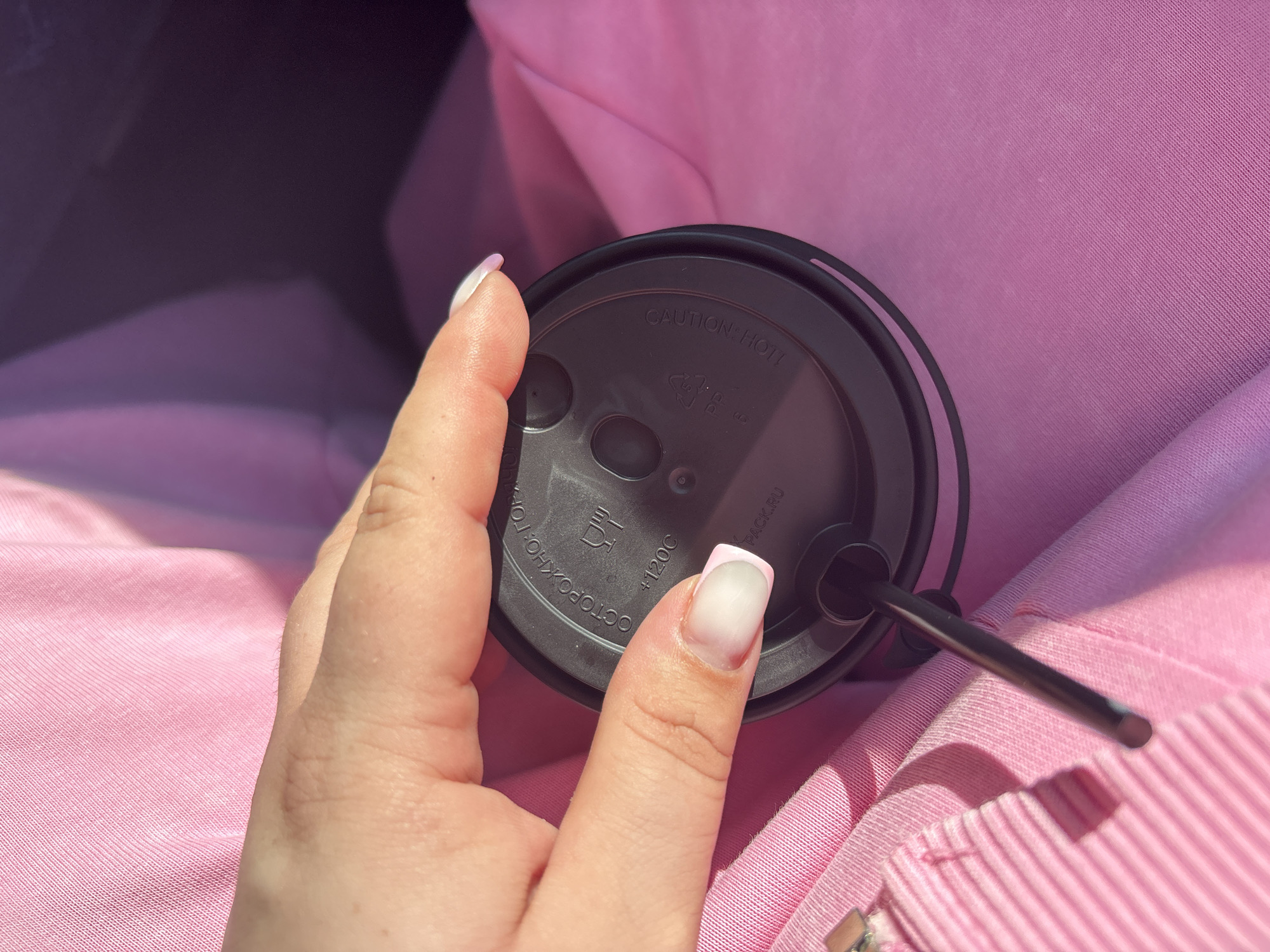 A close-up, top-down shot shows a person's hand holding a black lid of a disposable cup. The lid has text and symbols etched onto it. A black straw extends from the lid and rests on a pink fabric. The background is also pink fabric. No specific location in Вяйнянскі сельскі Савет, Belarus can be identified from this image.