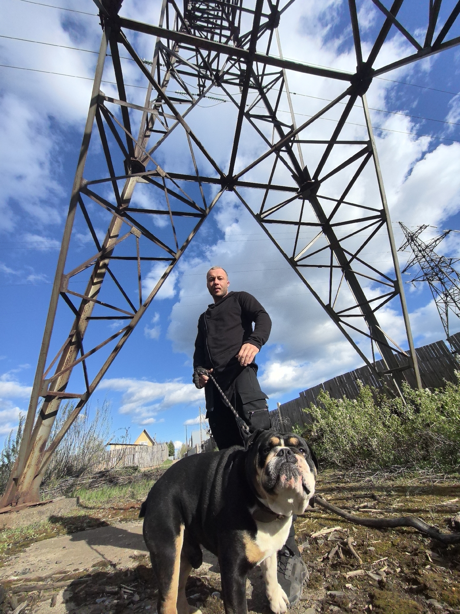 A medium shot shows a man standing with his bulldog underneath a metal electrical transmission tower against a bright blue, cloudy sky. The man is facing forward, wearing a black long-sleeved shirt and black pants, with a scowl on his face. He holds a black rope that is attached to the dog’s collar. The bulldog is black, tan, and white, with wrinkles on its face, and is looking up at the camera. The transmission tower is composed of metal beams arranged in a geometric pattern, with another tower visible in the background. The scene is located on a dirt and grass lot, with a wooden fence, shrubs, and a small shed visible in the background. The location is Noviy, Russia.