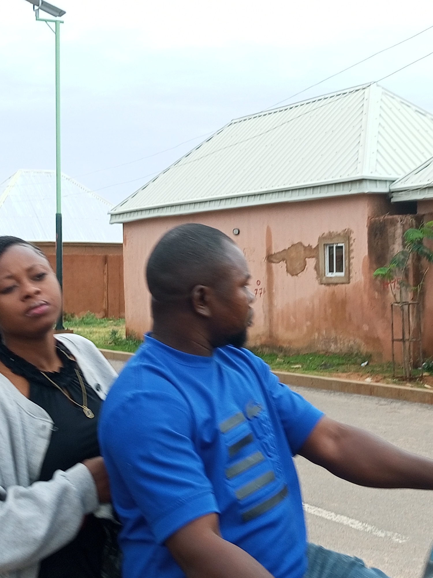 The image shows two people, a woman and a man, riding on a motorcycle in what appears to be a residential area of Gombe, Nigeria. The woman is in the foreground on the left, wearing a black top and a light gray cardigan, with a gold necklace. The man is on the right, wearing a blue t-shirt and blue jeans. He is operating the motorcycle.
In the background, there are buildings with light-colored metal roofs and reddish-brown walls. One building has a small white-framed window. There is a solar-powered street light to the left of the image. The ground appears to be asphalt, with some grass and dirt visible. The sky is overcast. The overall scene suggests a casual daytime ride in a semi-urban environment.