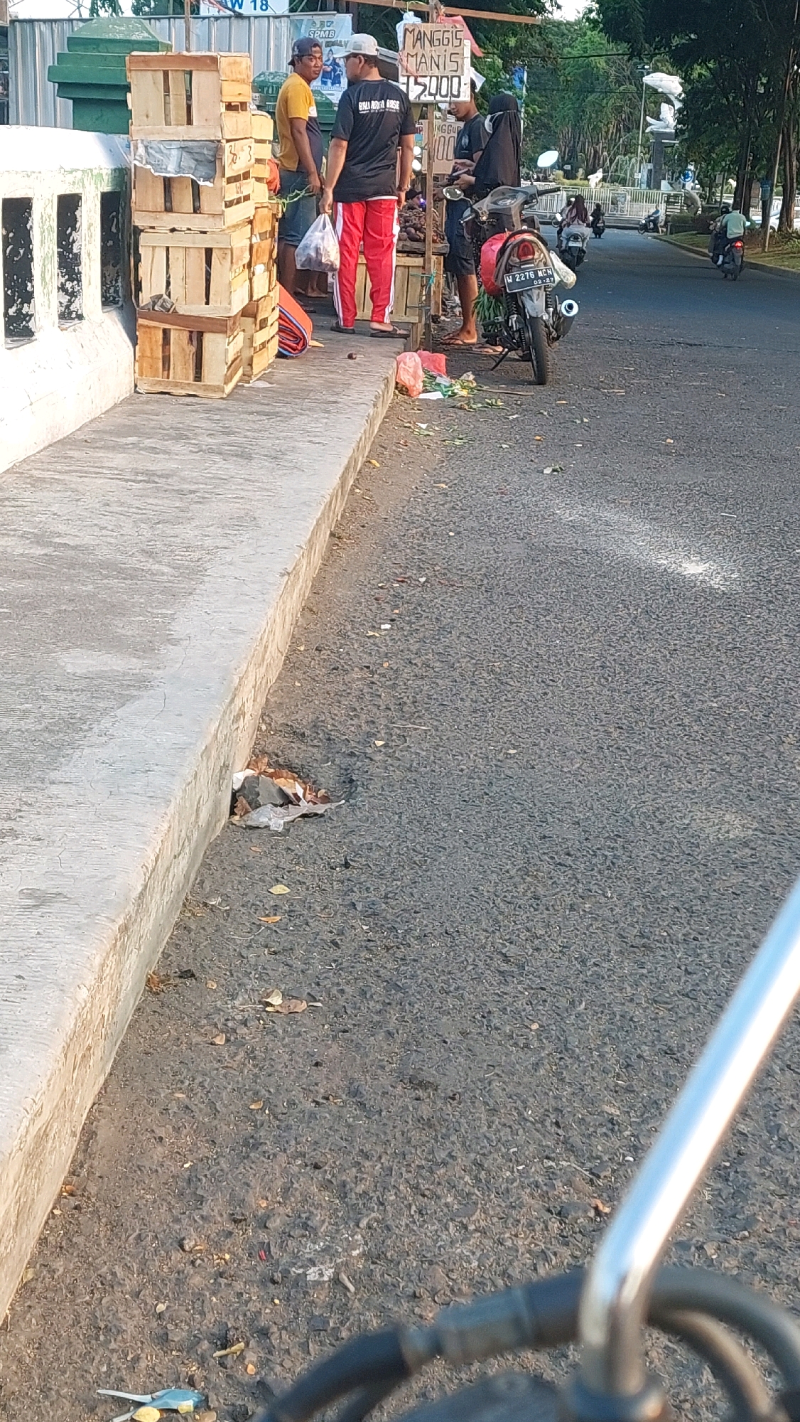 In Sepande, Indonesia, a street vendor is seen with a stack of wooden crates on a sidewalk next to a road. The vendor, wearing a black t-shirt and red pants, stands behind a stall displaying fruit for sale. Another individual, wearing a yellow t-shirt, is standing near the crates. Several motorcycles are parked and visible on the road in the background, with some people riding them. The scene depicts a typical street-side market environment with everyday activities.