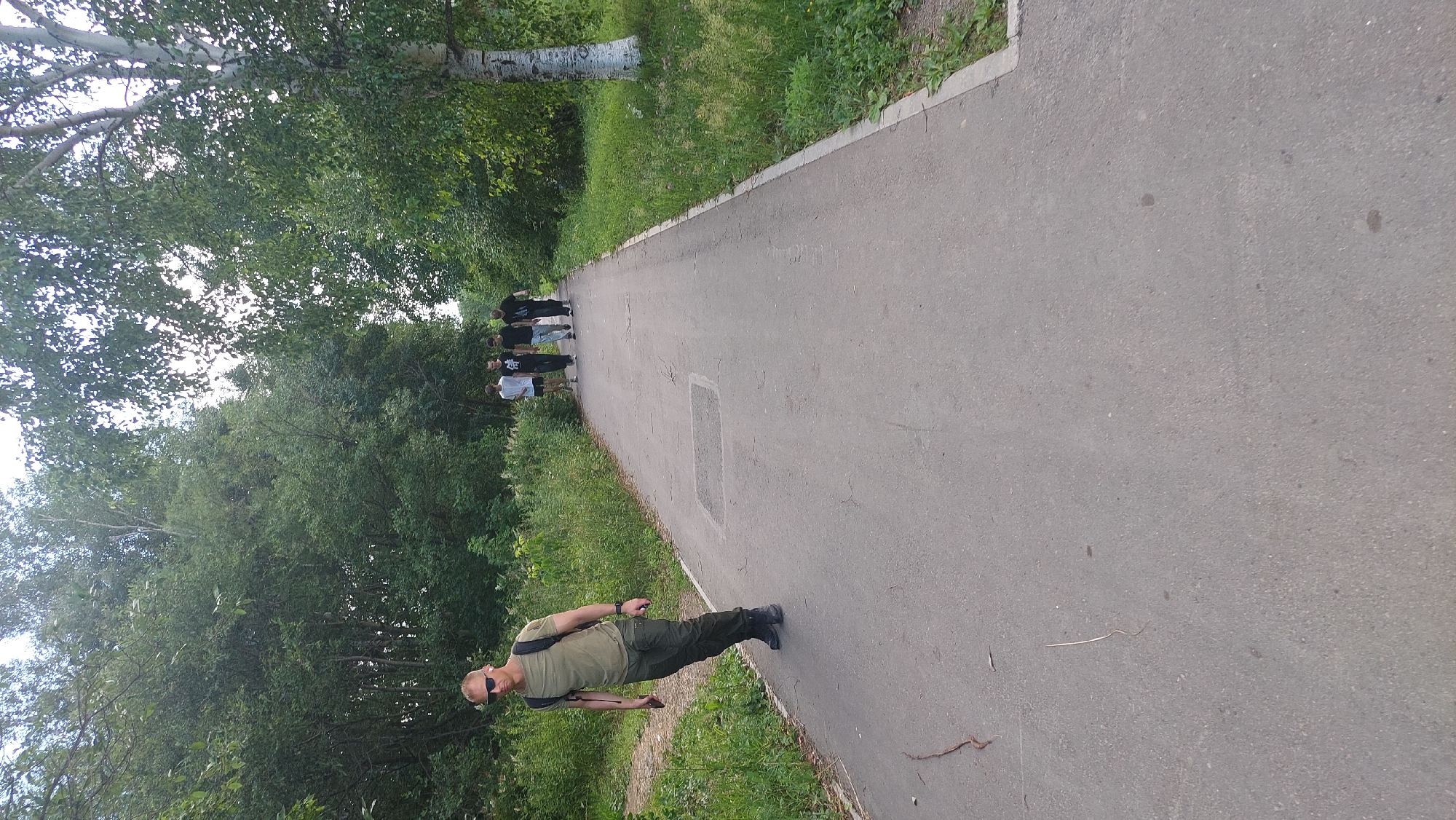 In Amursk, Russia, on a paved path bordered by lush greenery and mature trees, a man with short blonde hair, wearing sunglasses, a green t-shirt, and dark cargo pants, walks towards the camera with a backpack. He is holding a phone in his left hand. Behind him, a group of four young men walks away from the camera on the same path. The path is an asphalt surface with a visible square indentation. The environment is natural and serene, suggesting a park or a trail within the city. The lighting indicates it is daytime.