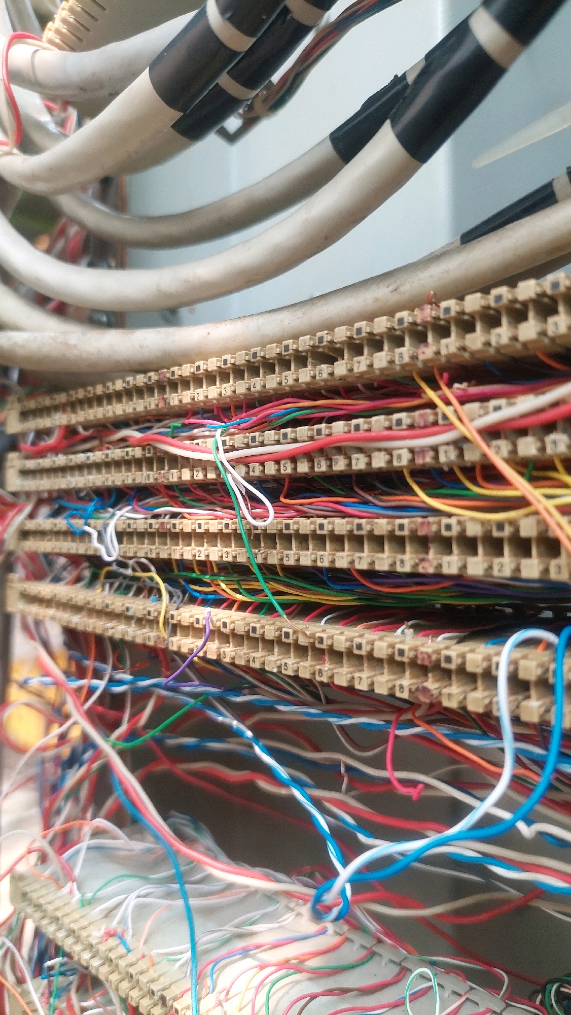 The image displays a close-up, eye-level view of a complex telecommunications wiring system, likely a distribution frame, within an indoor environment. Multiple rows of tan-colored connection blocks are visible, each containing numerous terminals numbered from 1 to 8. These blocks are densely packed with a multitude of colored wires, including red, blue, white, green, yellow, and orange, creating a tangled network. Some larger, white insulated cables are also present, bound together with black and white tape. The lighting suggests an artificial light source. There are no people or any discernible exterior environmental elements visible. The scene depicts organized, albeit messy, technological infrastructure. The location provided, Babakan Madang, Indonesia, is a town in West Java, known for its residential and agricultural areas, but this image focuses solely on the internal wiring.