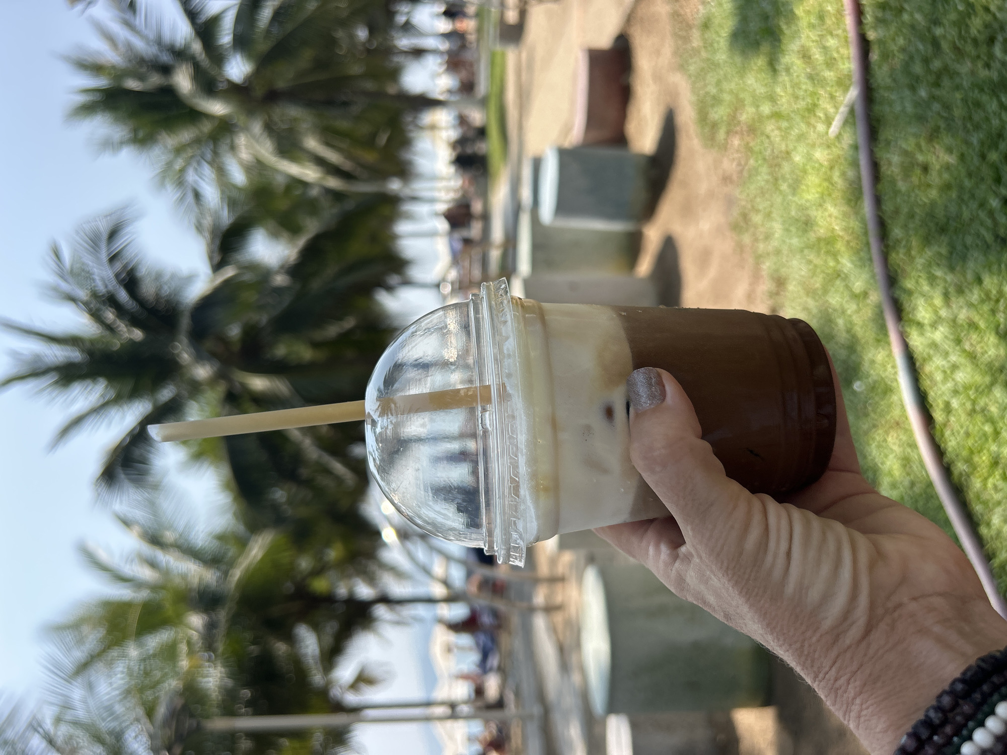 A hand holds a plastic cup filled with a beverage containing two distinct layers of brown and white liquids, topped with a clear dome lid and straw. In the background, there are out-of-focus palm trees and a blurry waterfront scene with people and structures, suggesting a coastal setting. The image was likely taken in Nha Trang, Vietnam, based on the visible scenery.