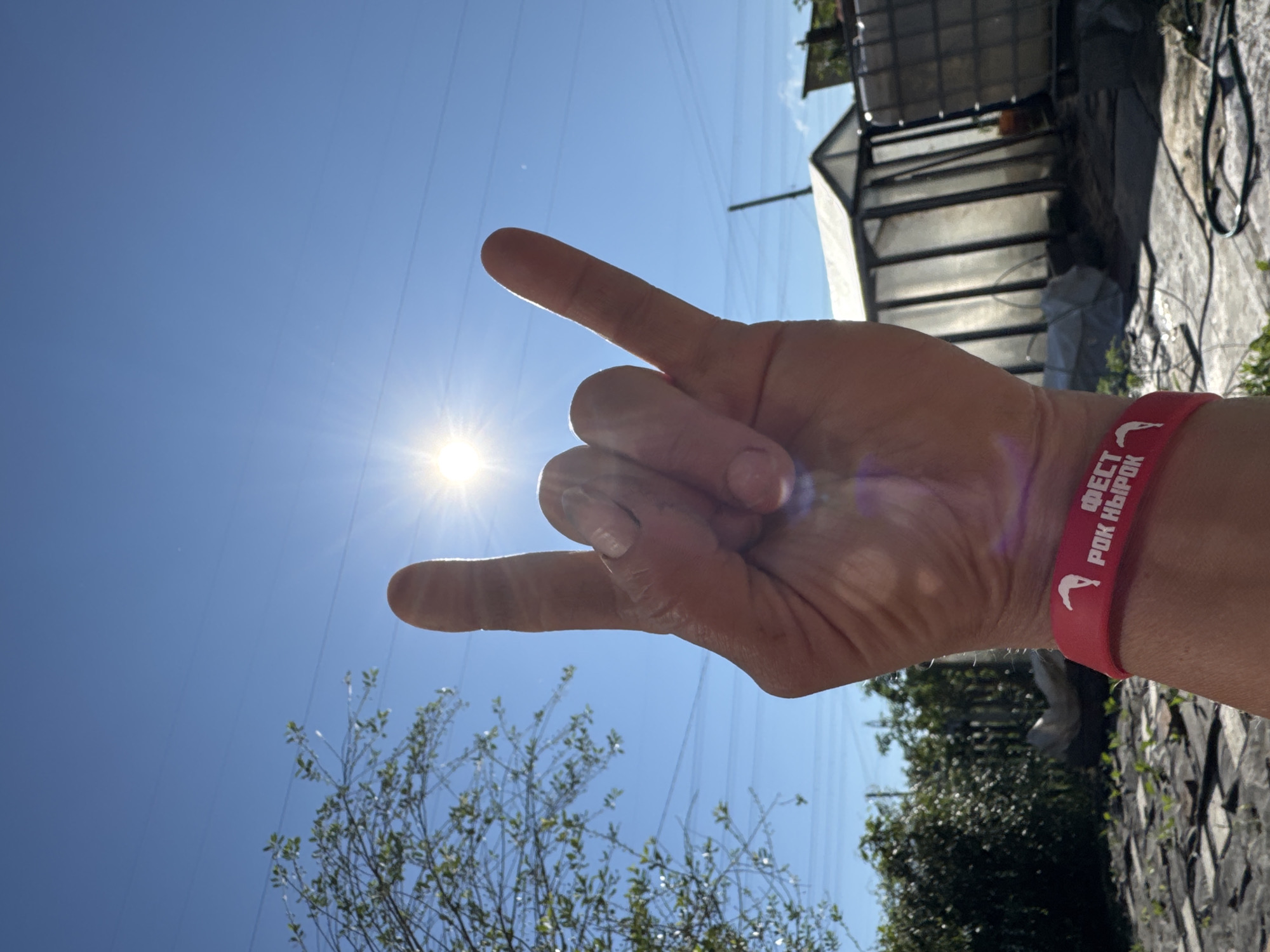 A person's hand forms the "rock on" gesture, with the index and pinky fingers extended, against a bright blue sky. The sun is visible, shining intensely with a lens flare effect. A red wristband with white text and a silhouette of a jumping figure is worn on the wrist. In the background, a tree with green leaves is partially visible on the left. Further in the background, a greenhouse-like structure with translucent panels and a large white water tank are present. The ground appears to be covered with dark fabric and some scattered debris. The image depicts an outdoor setting, possibly a yard or garden, under clear sunny conditions. This scene could be located in Chaikovsky, Russia, given the provided context.