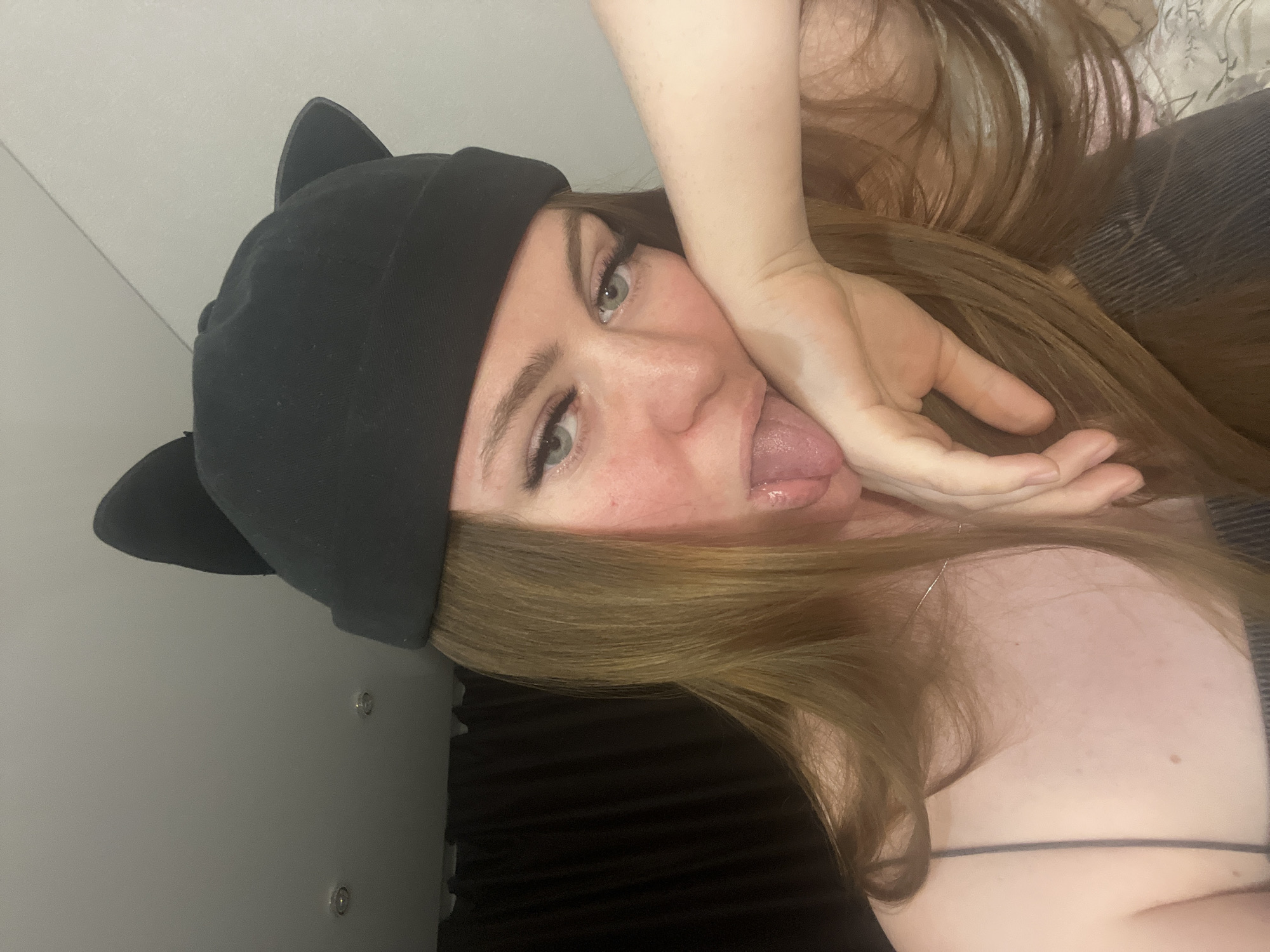 A fair-skinned individual with long, light brown hair, wearing a black beanie with cat ears, is shown with their tongue extended. One hand is held to their face. They are wearing a top with thin straps. The background consists of a light gray wall with two screw-like fasteners and a dark, textured surface. The location in the city of Sevastopol, Ukraine cannot be determined from the provided image.