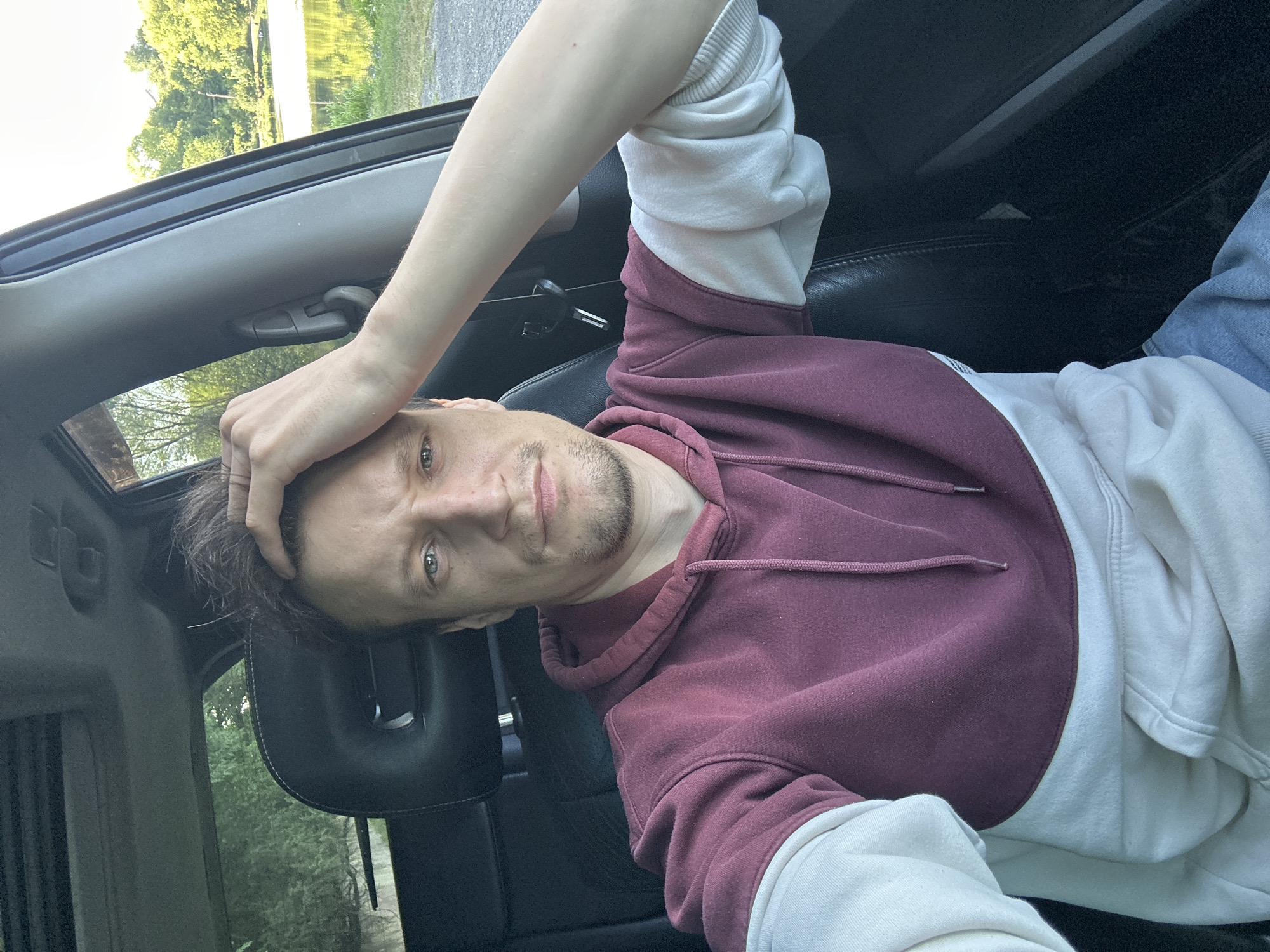 A young Caucasian male with light skin, short brown hair, and blue eyes is seated in the driver's seat of a car. He is wearing a maroon and cream-colored hoodie. His right arm is raised, with his hand resting on his head, and his left arm is resting on the car's interior. His expression is neutral, with his gaze directed forward. The car's interior is dark, with black leather seats and a sunroof visible above. Through the open car window, a body of water and a lush green treeline can be seen, suggesting an outdoor, possibly rural, setting. There is no visible indication that this scene is in Makiv, Ukraine.