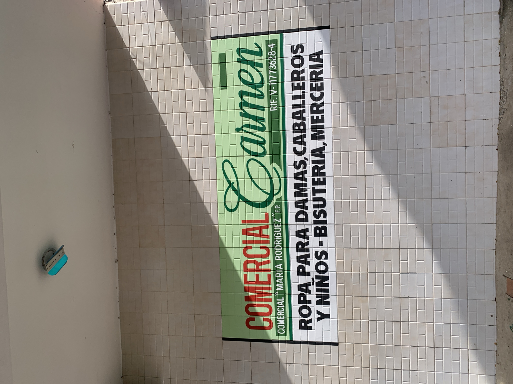 This outdoor shot captures a tiled wall featuring a prominent business sign, likely of a commercial establishment in Tacarigua, Venezuela. The wall is covered in light-colored, rectangular tiles. A strong, diagonal shadow is cast across the scene from the upper left to the lower right, indicating a bright, sunny day, likely mid-day or afternoon.
The main subject is a large, rectangular sign affixed to the tiled wall. The sign is designed with a green background on the left side, featuring the business name "COMERCIAL Carmen" in red and green lettering, respectively. Below "COMERCIAL," smaller green text reads "COMERCIAL 'MARIA RODRIGUEZ' F.P." A vertically oriented text block to the right of "Carmen" displays the Venezuelan tax identification number: "RIF. V-11773628-4".
The right side of the sign has a white background with bold black text, detailing the types of products offered: "ROPA PARA DAMAS, CABALLEROS Y NIÑOS - BISUTERIA, MERCERIA," which translates to "Clothing for ladies, gentlemen, and children - costume jewelry, haberdashery."
To the left of the tiled wall, a plain light-colored wall is visible, on which a small, bright blue object is mounted on a metallic bracket. Its specific function is unclear, but it appears to be either reflective or illuminated. No people or direct activity are depicted; the image focuses on the static architectural elements and the business signage.