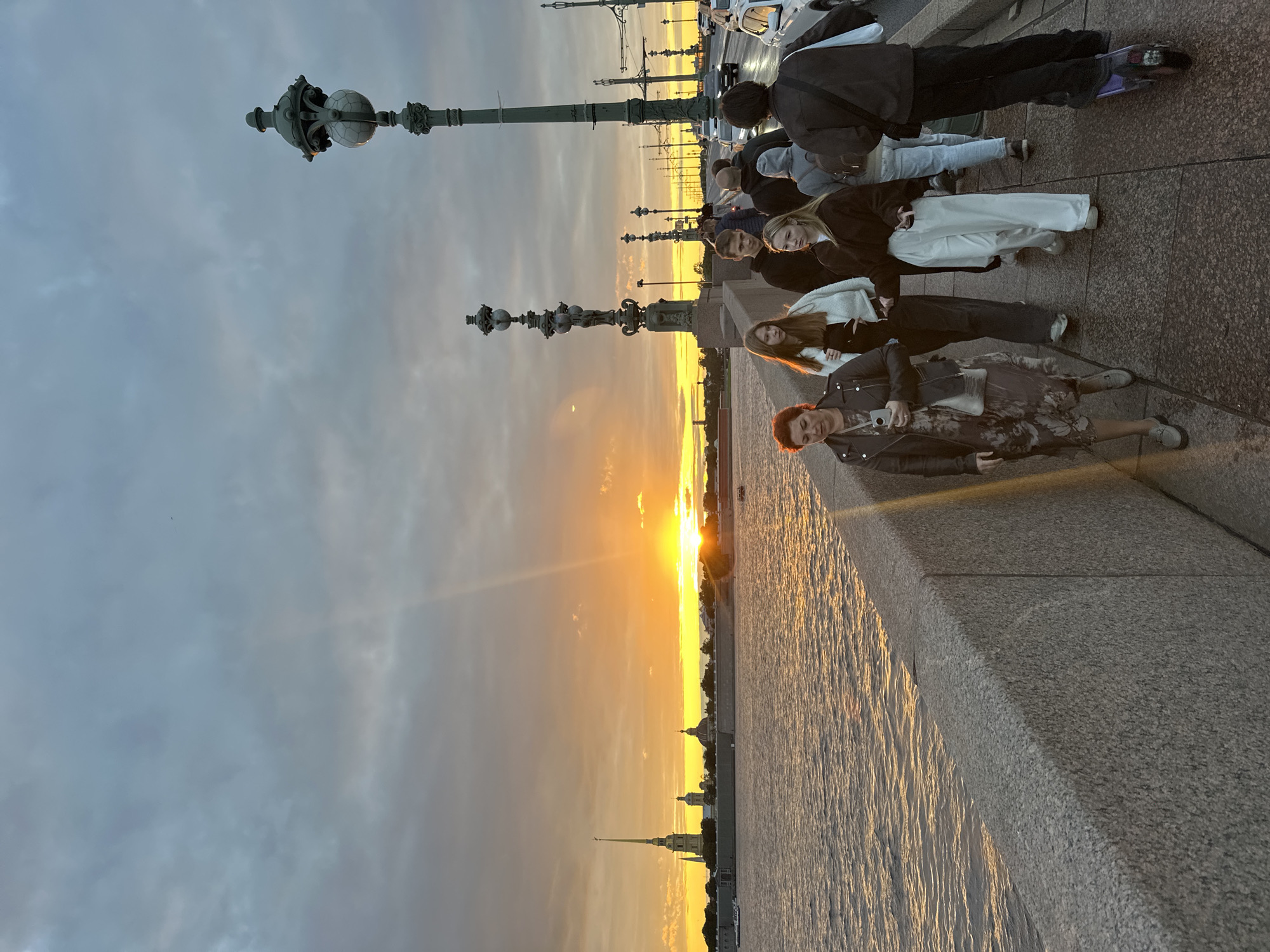 The image depicts a sunset over the Neva River in Saint Petersburg, Russia, viewed from the Palace Bridge. The sky is filled with a dramatic sunset, with hues of orange and yellow radiating from the horizon, casting a warm glow on the water. In the distance, the Peter and Paul Fortress is visible, its spire piercing the twilight sky. A group of people are walking across the bridge, some strolling leisurely, while others are seen on an electric scooter. The bridge itself is lined with ornate lampposts, characteristic of the city's architecture. Cars are also present on the bridge, indicating its role as a transportation route. The overall atmosphere is one of a serene evening in the city, with the natural beauty of the sunset juxtaposed with urban activity.