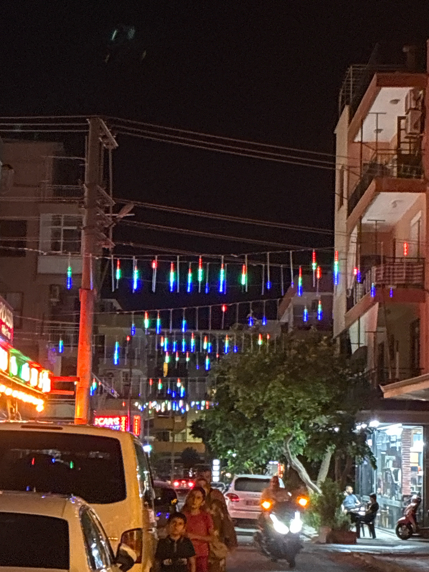 This is a nighttime photograph taken in Alanya, Turkey, likely on a street with shops and residential buildings. Colorful string lights are hung across the street, creating a festive atmosphere. Several people are visible in the image: a group of individuals, including children and adults, walking on the sidewalk; a person seated at a table outside a shop; and a person riding a scooter. Vehicles, including a white van and a white SUV, are parked or driving on the street. The buildings appear to be multi-story structures with balconies. The overall scene suggests a lively urban environment at night.