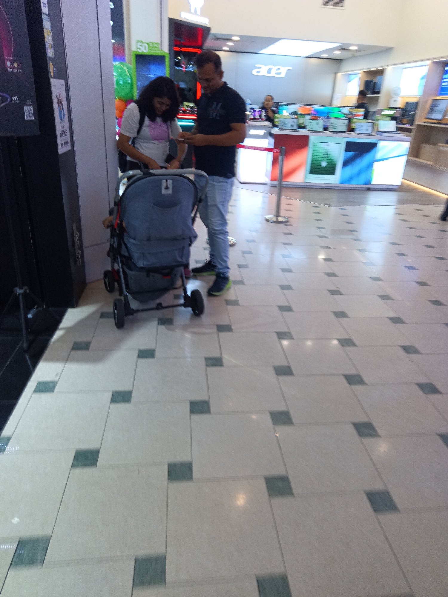 An Asian man and woman are standing in front of a stroller in a retail store. The man is wearing a black t-shirt and blue jeans, and the woman is wearing a white t-shirt and dark pants. The man appears to be looking at his phone. The stroller is grey and has a child in it, though the child is not visible. In the background, there is a display of Acer products, including laptops and monitors. The store is well-lit with fluorescent lighting, and the floor is tiled with a diamond pattern of white and green tiles. The store appears to be located in Kuala Lumpur, Malaysia, based on the signage and overall aesthetic of a modern shopping mall.