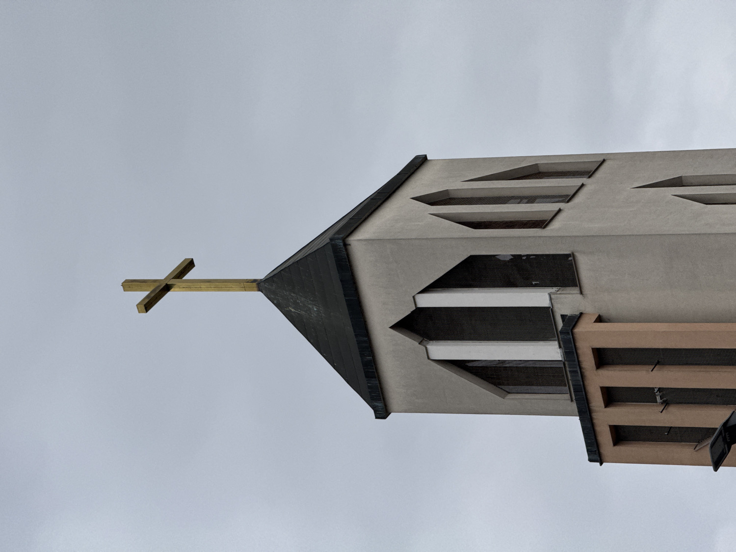 The image captures an upward-angled view of the upper section of a modern church or religious building, set against a uniform, overcast grey sky. A prominent golden cross is fixed at the peak of the main structure, extending from a dark, angular roofline.
The building itself is constructed from a light grey material, possibly concrete or stucco, and features a distinctive modern architectural style characterized by sharp angles and geometric shapes. Several deeply recessed windows or vents are visible, covered with dark grates. On the main grey facade, these openings are elongated parallelograms, arranged horizontally. An adjoining section, likely a tower or an extension, is visible to the right, rendered in a contrasting beige or light brown color. This section also incorporates similar grated openings, arranged vertically.
The sky is a pale, uniform grey, indicating an overcast daytime. No people or activities are visible. The overall aesthetic is minimalist and structural, highlighting the angular forms and material contrasts of the building. The scene is situated in the City of Zagreb, Croatia. There is no visible text in the image.
