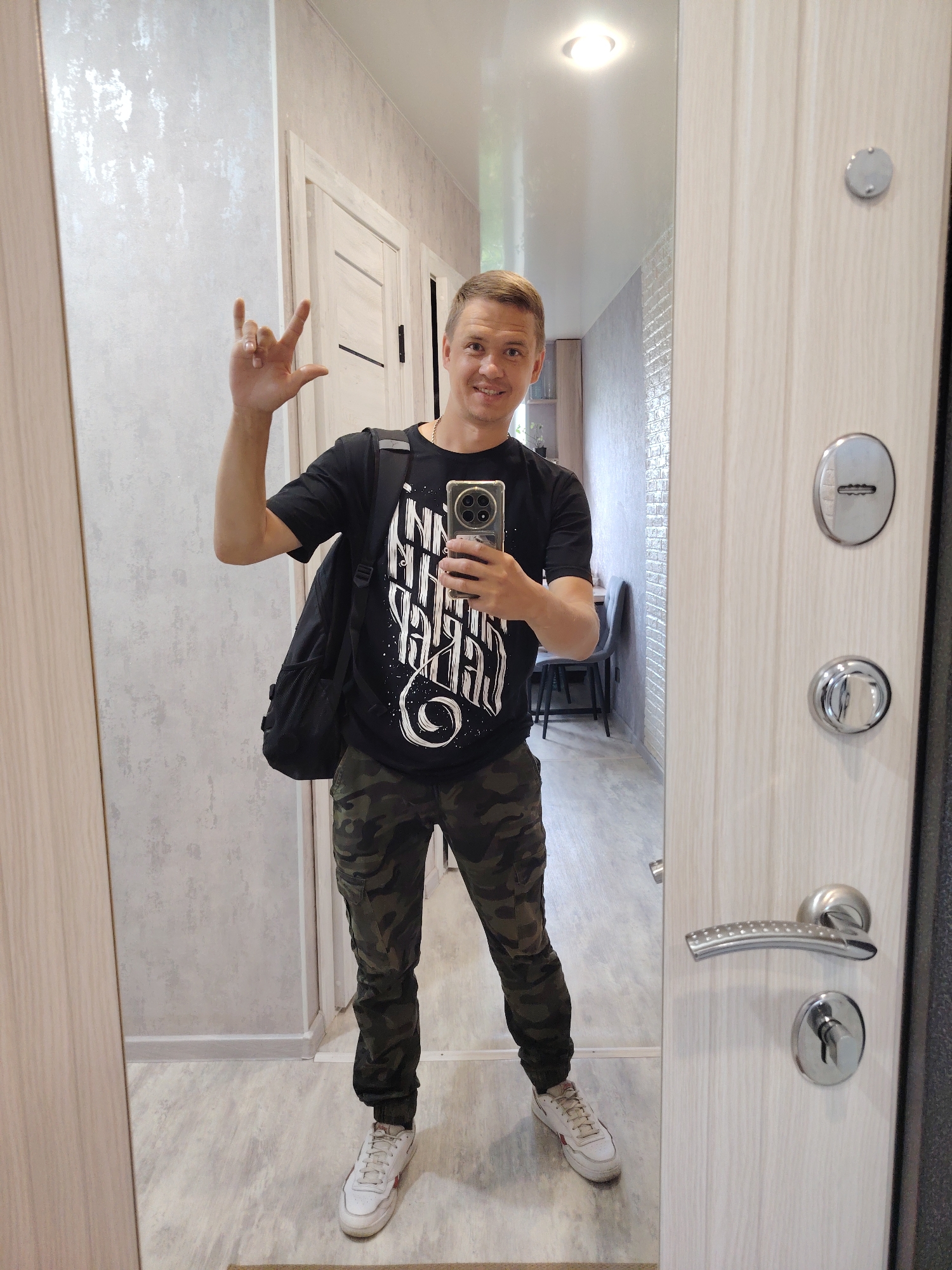 A young man is standing in front of a mirror, taking a selfie. He is wearing a black t-shirt with white text, camouflage pants, and white sneakers. He has a black backpack slung over his left shoulder and is holding a smartphone in his right hand. He is making a "rock on" hand gesture with his left hand. The mirror reflects a hallway with light-colored walls and doors. The man is in a residential building. The image is a selfie taken in the city of Chaikovsky, Russia.