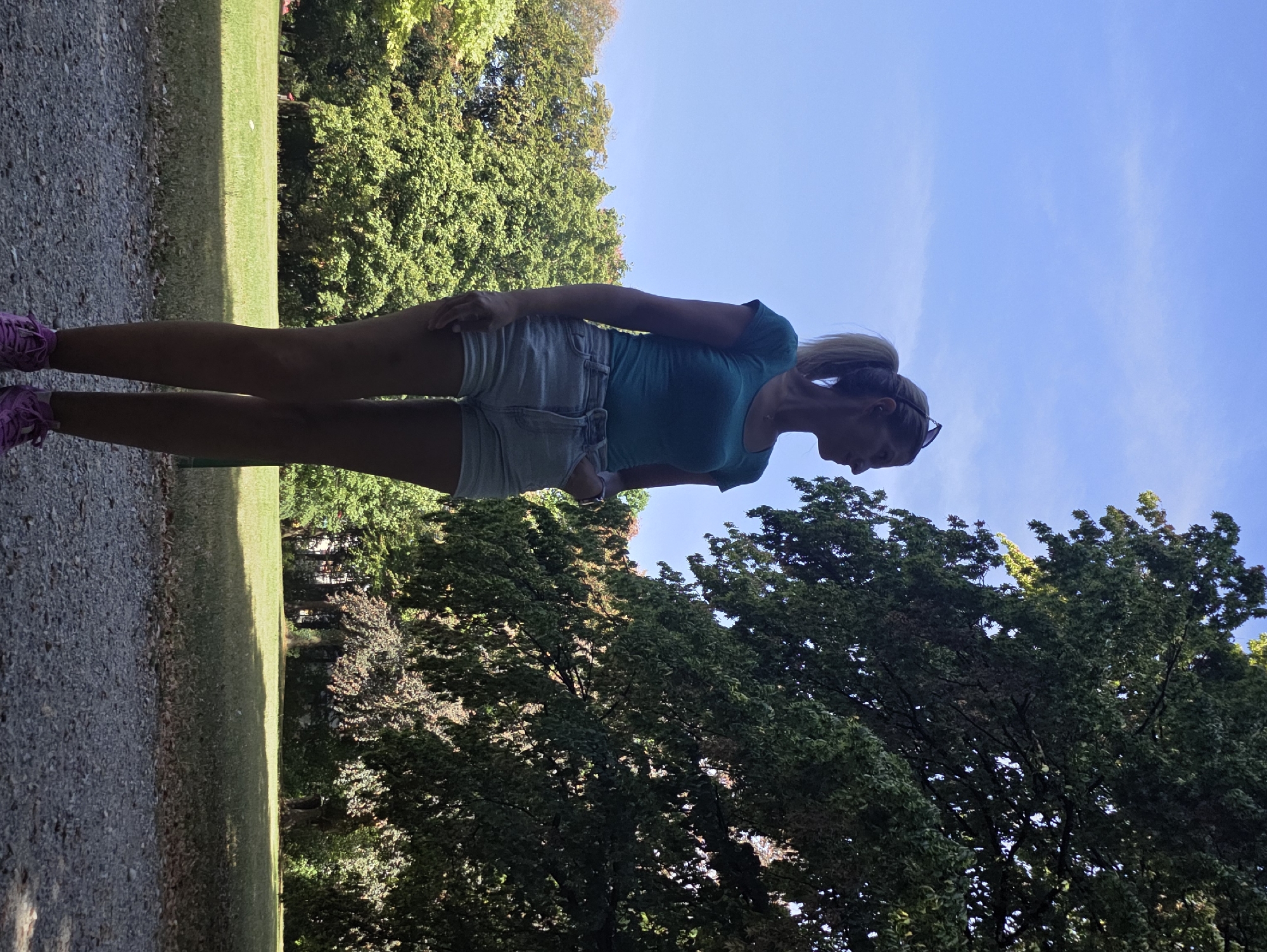 A woman is standing in a park, looking down at the ground. She has blonde hair pulled back in a ponytail and is wearing a turquoise t-shirt and light-colored denim shorts. She is also wearing pink athletic shoes. The background consists of lush green trees and a clear blue sky. The sun is shining, indicating it is daytime, and the weather appears to be pleasant. The setting is a park, likely in Munich, Germany, given the context. The woman's posture suggests she might be pausing for a break or observing something on the ground.
