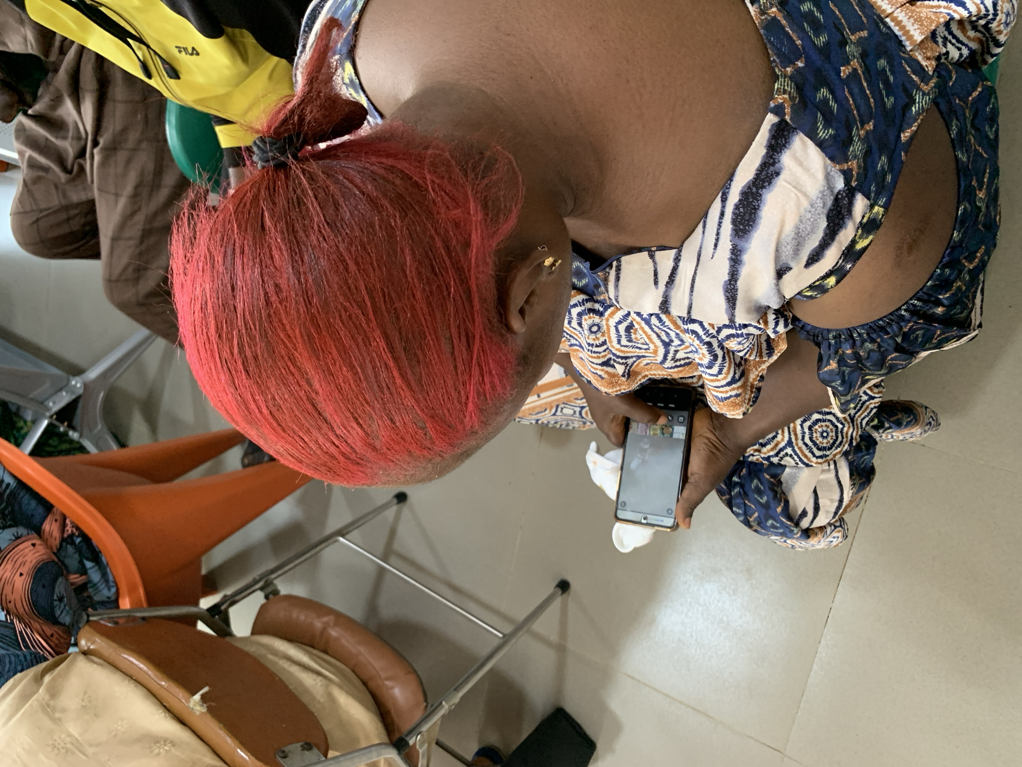 A person with bright red hair is seen from the back, holding a smartphone. The person is wearing a patterned top. The background shows a room with tiled flooring and several chairs, one of which is bright orange. The scene appears to be indoors. No specific location in Jos, Nigeria is identifiable from the image.