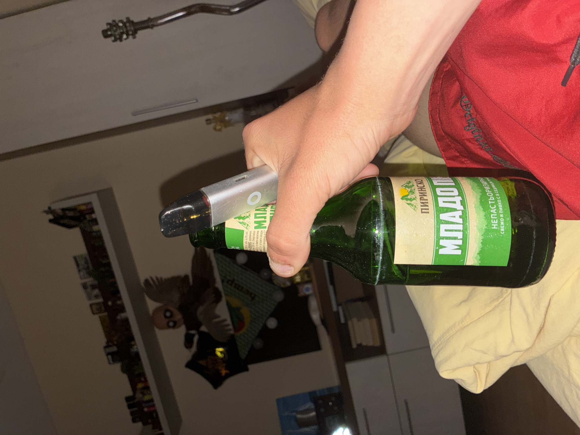 The image captures a close-up, slightly angled view of a person's hand holding a green glass bottle and a silver electronic device indoors. The person wears red shorts and possibly a light-colored top, with their lower arm and hand prominently featured.
The main subjects are the person's hand, the bottle, and the electronic device. The device appears to be a vape pen, featuring a circular button and a sleek, metallic design. The bottle is a beer bottle with a cream and green label. Visible Bulgarian text on the label includes "ПИРИНСКО МЛАДО ПИВО" (Pirinjsko Mlado Pivo - Pirinjsko Young Beer), indicating it's a Bulgarian brand. Further text reads "НЕПАСТЬОРИЗИРАНО" (Unpasteurized) and "СВЕЖО И ПИВКО С 8 СЕДМИЦИ ПРО" (Fresh and drinkable for 8 weeks), along with "4.4% vol." The word "МЛАДО" (Mlado - Young) is prominently displayed.
The setting is an indoor room, likely a bedroom or casual living space in Varna, Bulgaria, given the linguistic cues on the bottle and the provided context. In the blurred background, various objects are visible, including a shelf or cabinet filled with items, a dark, plush-like object resembling a stuffed animal (possibly a deer head), and another pillow or bag with the faint word "hemp" on it. A metal fixture is visible on the ceiling. The lighting suggests an artificial light source, possibly indicating evening or nighttime. The activity depicted is simply the person holding both the bottle and the electronic device.