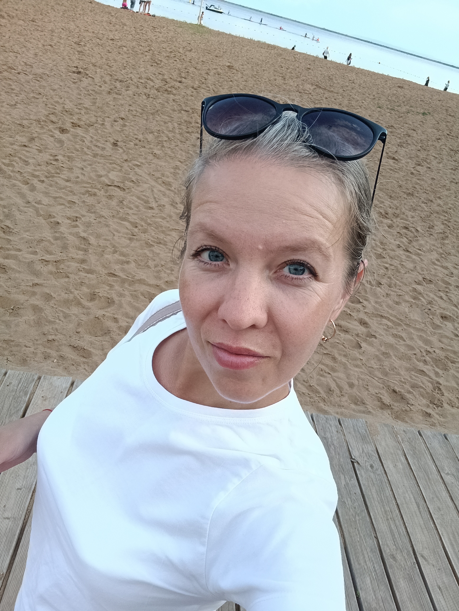 A woman with graying blonde hair and blue eyes takes a selfie on a wooden boardwalk at a beach. She is wearing a white t-shirt and has sunglasses perched on her head. The beach is sandy, and in the background, several people are visible near the water, some wading or standing, while others appear to be on a boat or paddleboard. The sky is overcast, and the water is a muted blue. The scene is likely at the Kama River embankment in Chaikovsky, Russia, a popular recreational area.