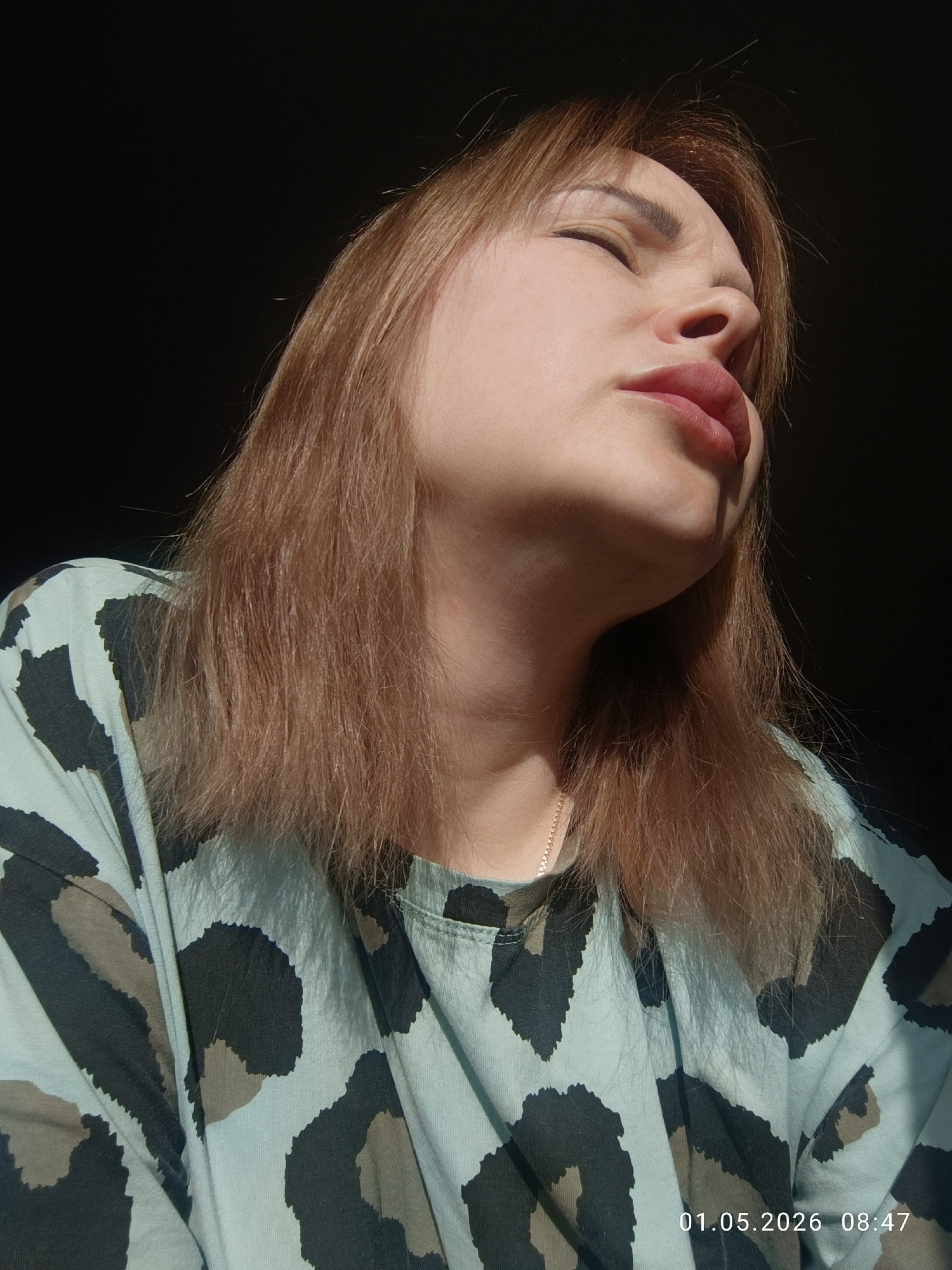 A woman with fair skin and light brown hair stands in a dimly lit indoor space. She is wearing a pale blue animal print shirt. The subject is looking up and slightly to the side, her eyes are closed and her face is tilted. A thin gold chain necklace is visible around her neck. Parts of her face and shirt are illuminated by an unseen light source. The background is dark. A date of 01.05.2026 and time of 08:47 are printed on the bottom right. While there is no information in the photo to definitively locate this scene in Chaikovsky, Russia, the date and time are provided as a visual element in the lower right corner.