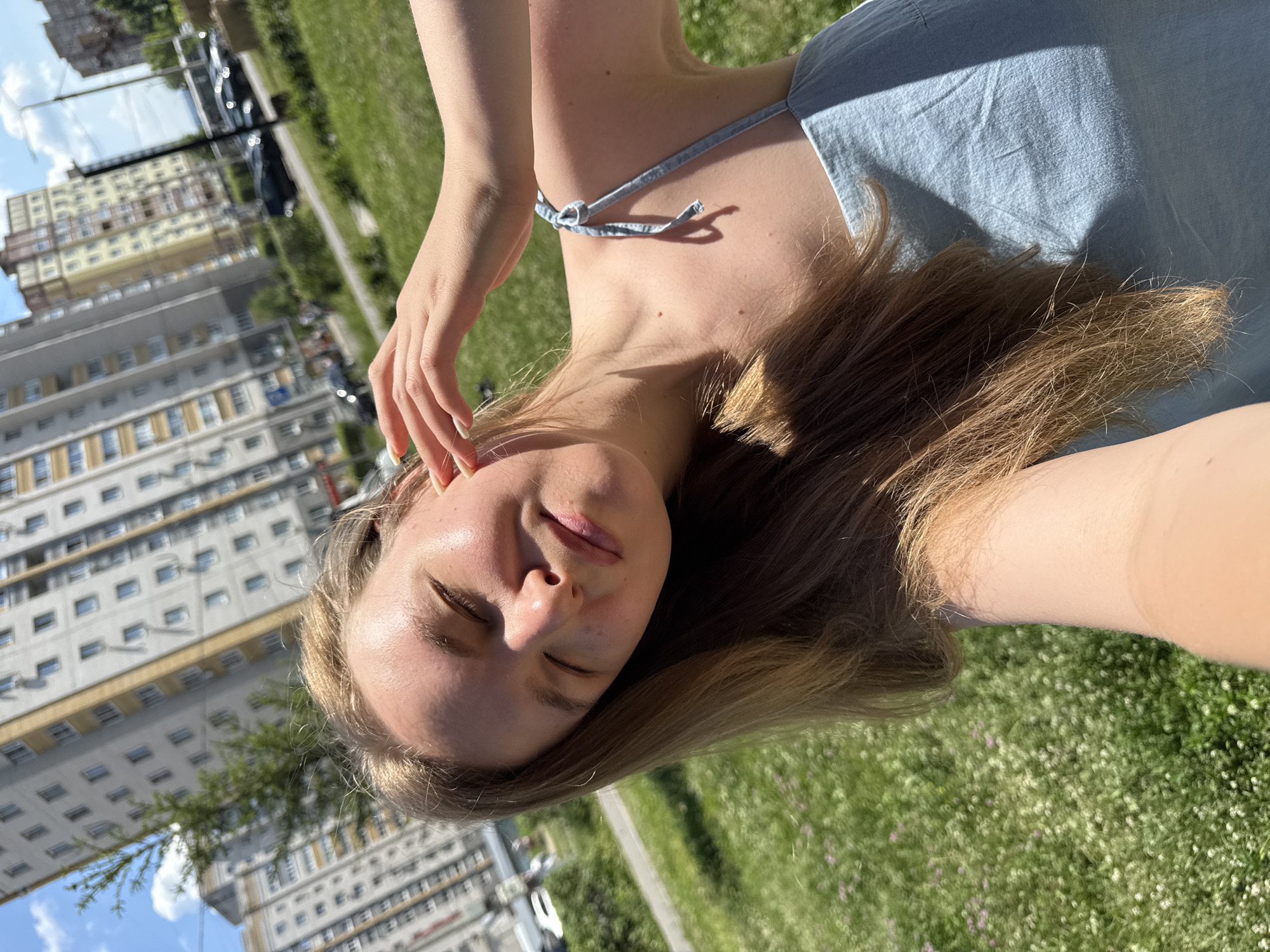 A young woman with blonde hair is taking a selfie outdoors in the city of Vladimir, Russia. She is wearing a light blue denim sundress. Her eyes are closed, and her hand is resting on her head. The background shows apartment buildings, a street with cars, and a grassy area with small flowers. The sun is shining brightly, casting shadows on the woman's face and shoulder. The overall scene suggests a warm, sunny day in an urban environment.