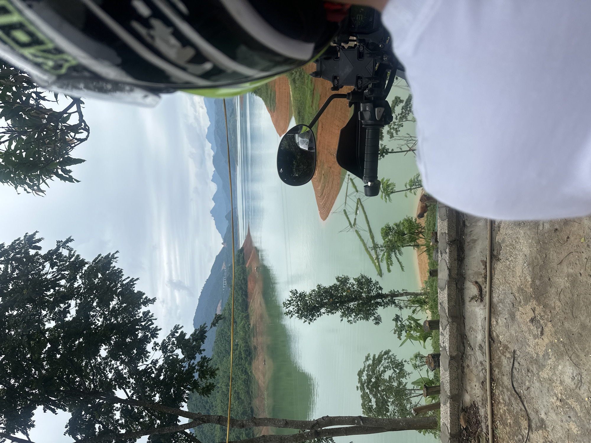 In Bản Phúc, Vietnam, a person wearing a white t-shirt and a black and green helmet is sitting on a motorcycle. The motorcycle's handlebars and a rearview mirror are visible in the foreground, reflecting a glimpse of trees and the sky. The scene looks out onto a serene lake with calm, turquoise water. The lake is bordered by lush green hills and mountains in the distance, some of which are shrouded in mist. The shoreline exhibits exposed, reddish-brown earth, hinting at a receding water level or natural erosion. Scattered trees and vegetation dot the landscape around the lake. The sky is overcast, suggesting a cloudy day.
