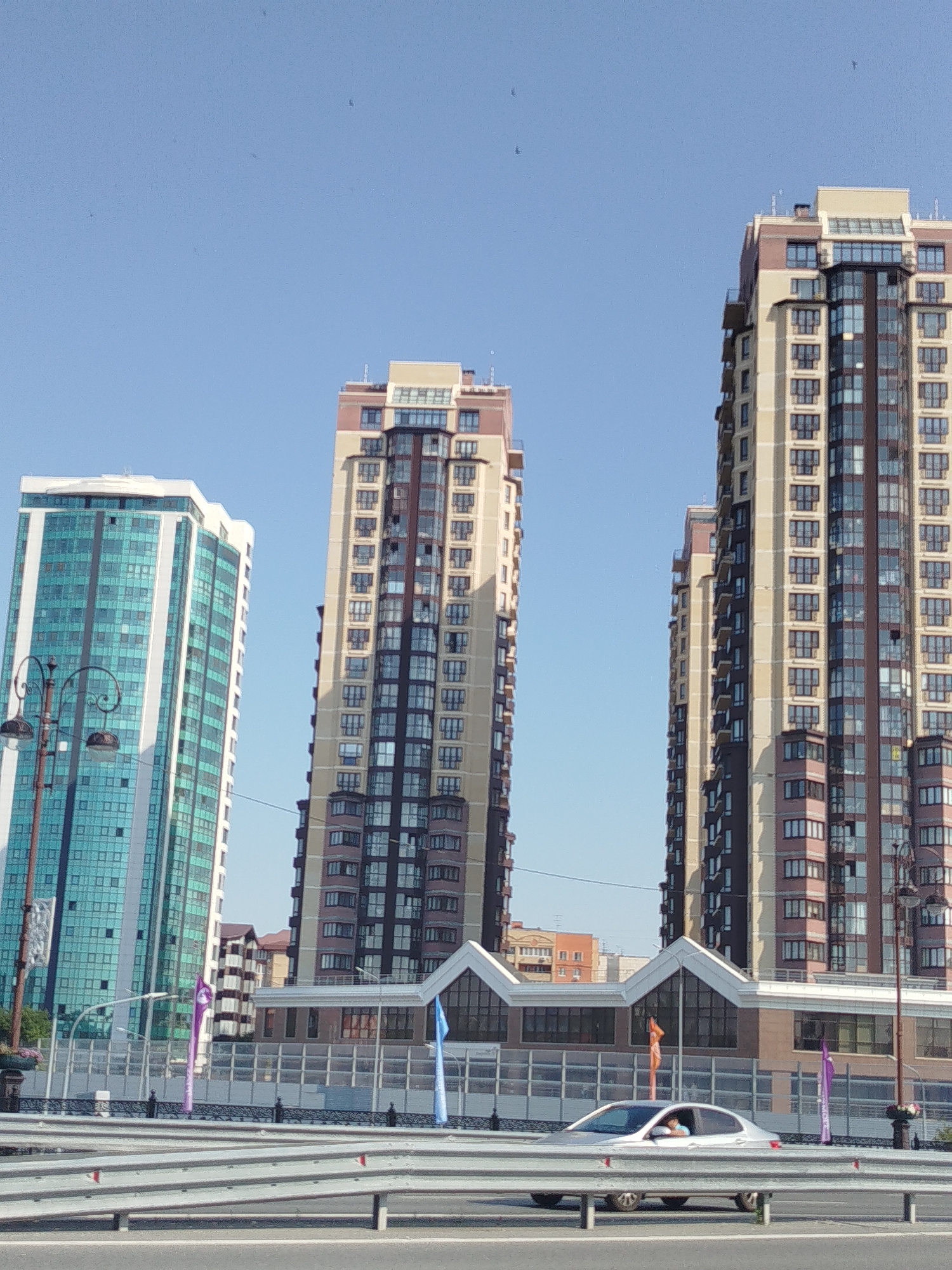 This is a photograph taken in Tyumen, Russia, showing a view of several modern high-rise apartment buildings against a clear blue sky. The building on the left is predominantly glass with a teal tint and white structural elements. To its right are two taller buildings, one with a beige and brown facade and numerous balconies, and another similar building slightly behind it. In the foreground, a silver car is visible driving on a road behind a guardrail. The car has one person visible in the driver's seat. Flags of different colors, including purple and blue, are visible on flagpoles between the buildings and the road. A decorative lamppost is also present on the left side of the image. The scene appears to be during the daytime.