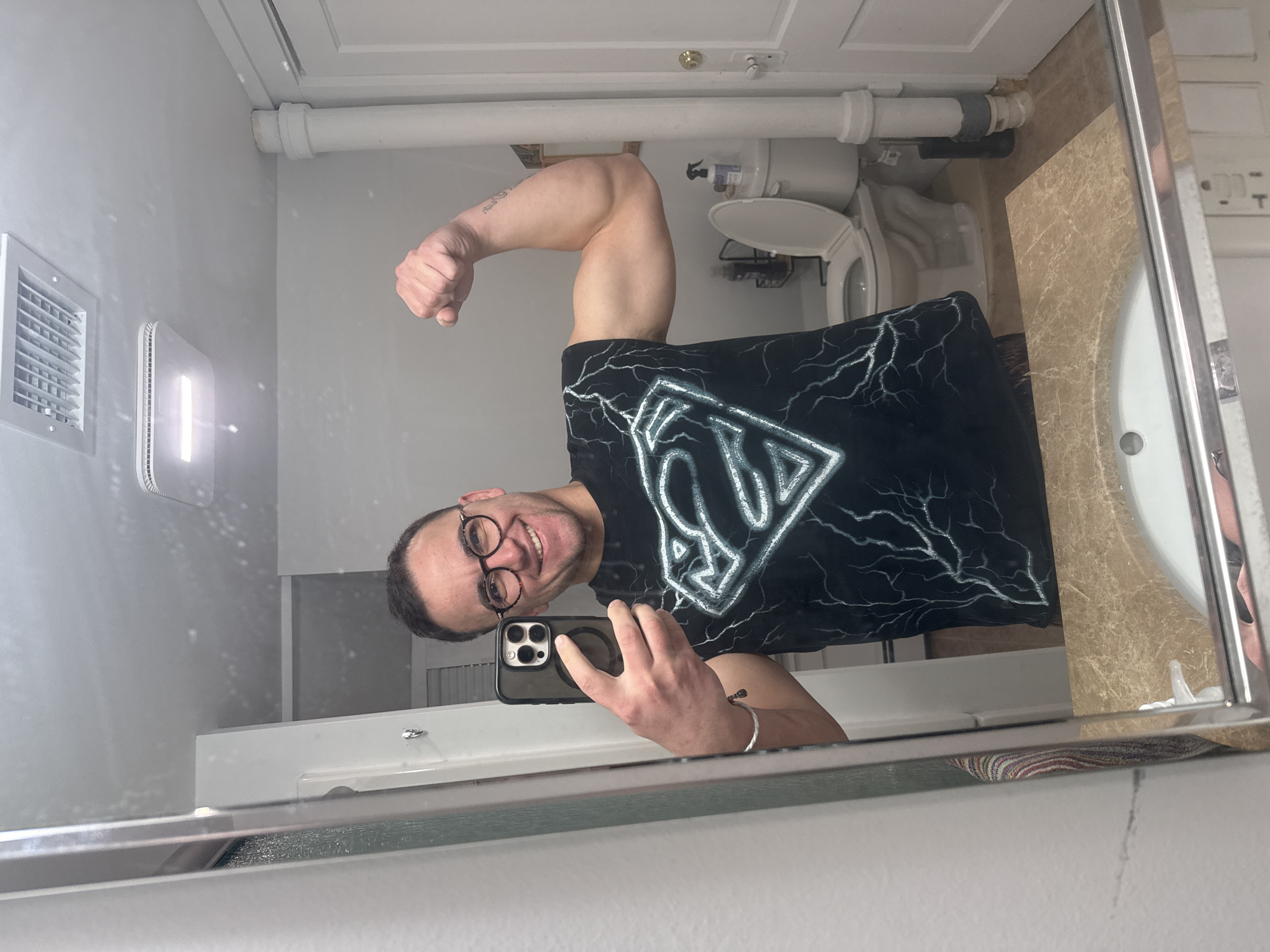 A man with short dark hair and glasses is taking a mirror selfie in a bathroom. He is wearing a black t-shirt with a stylized Superman logo and lightning designs. He is flexing his right bicep. The bathroom includes a toilet, sink with a beige countertop, a ventilation grille, and a pipe running along the ceiling. The location cannot be verified as Provincetown, United States of America.