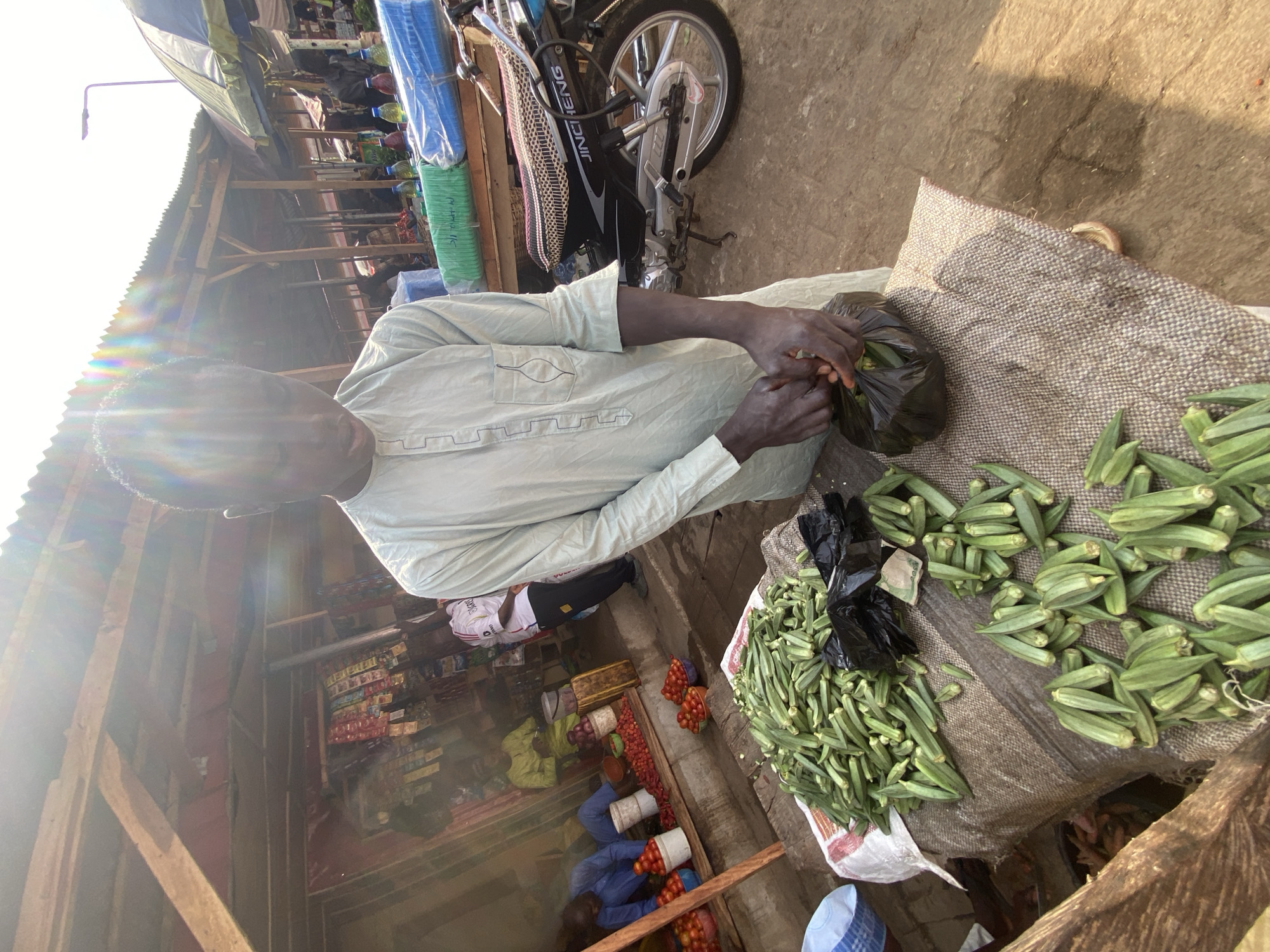 This image captures a vibrant, sunlit scene at an outdoor market in Kafanchan B, Nigeria. The central figure is a man, likely a vendor, wearing a light-colored, long-sleeved garment. He is focused on handling fresh green okra, which he appears to be placing into a black plastic bag. Several large piles of okra are spread out on a coarse, woven mat beside him, ready for sale.
Behind the vendor, a silver and black motorbike is parked, featuring the visible text "JINCHENG" on its side. The background reveals a lively market environment with simple, open-air stalls constructed from wooden frames and corrugated iron roofs. Bright sunlight causes significant lens flare across the top left and center of the image, indicating either morning or late afternoon.
Further into the market, other individuals are visible, some kneeling or sitting near their own produce displays, including mounds of red tomatoes and onions. Shelves stocked with various packaged goods can be seen in the distance, underscoring the commercial nature of the setting. The ground is a mix of dirt and concrete. The scene conveys the busy, communal atmosphere of a typical West African market, bustling with daily commerce under clear, sunny skies.