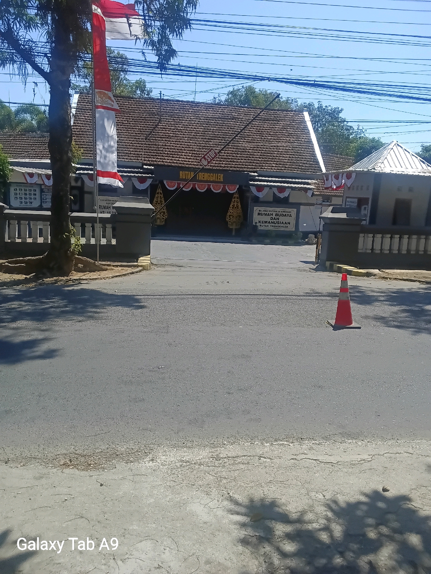 The image shows the entrance to a government building in Trenggalek, Indonesia, identified by the sign that reads "RUTAN TRENGGALEK". The building has a tiled roof and is adorned with red and white banners, a national flag, and traditional Javanese puppets. A traffic cone is visible on the asphalt road in front of the building. The sky is clear and blue, with power lines crisscrossing overhead. Trees and shadows are also present in the scene. The overall impression is one of a public institution on a sunny day, possibly during a national holiday or event, given the festive decorations.