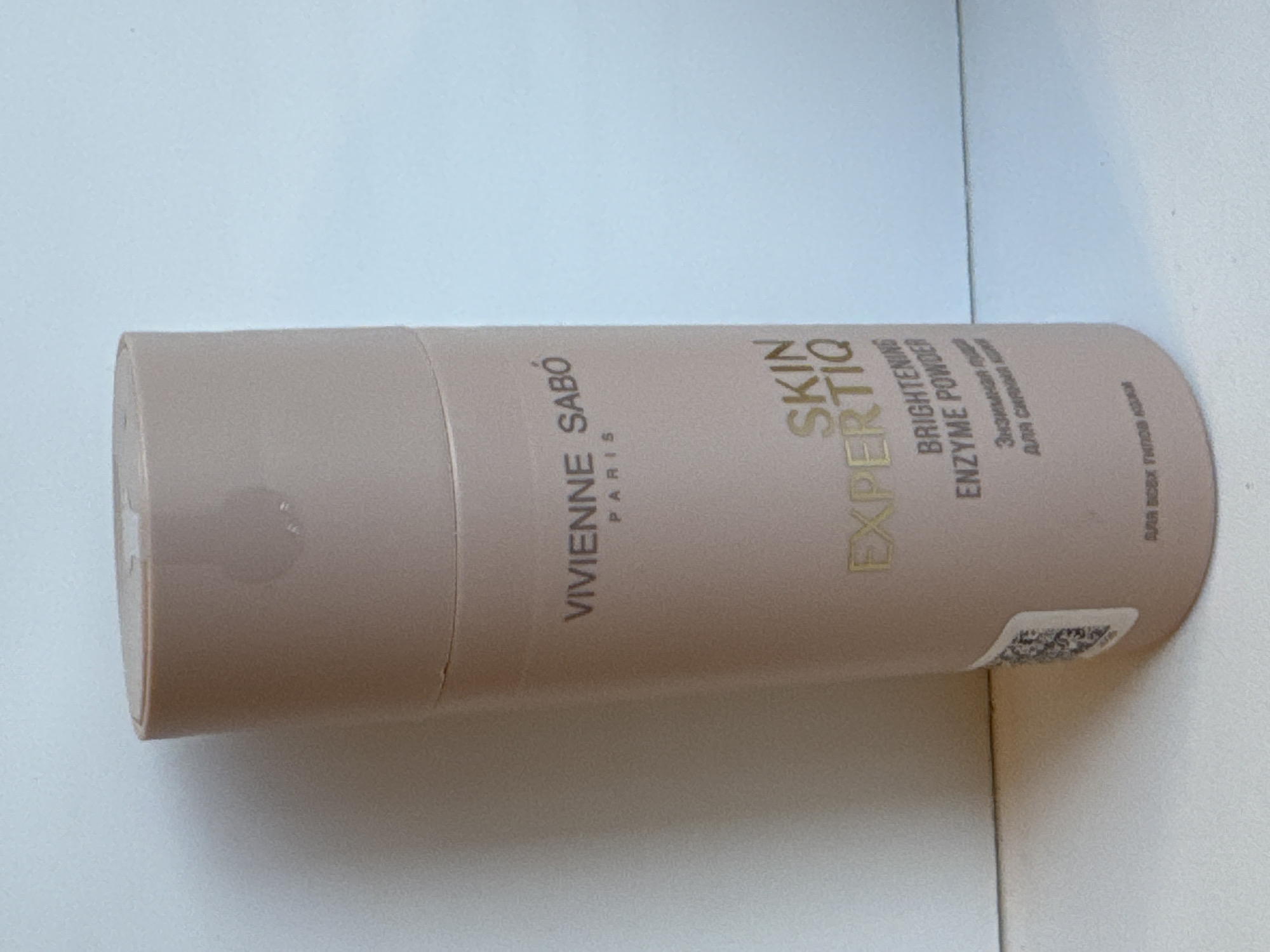The media file displays a close-up, side-view of a cylindrical cosmetic product, specifically a "Vivienne Sabo Skin Expertiq Brightening Enzyme Powder." The product is a light, neutral-toned tube, lying horizontally on a white surface, likely a tabletop. The lighting suggests an indoor environment, possibly a domestic setting, with no distinct time of day or weather cues.
The tube features prominent gold lettering. The brand name "VIVIENNE SABO" and "PARIS" are clearly visible. The product name "SKIN EXPERTIQ" is also legible, followed by "BRIGHTENING ENZYME POWDER." Below this, in smaller text, is "Энзимная пруд" and "DRA CARINA" which appear to be in Cyrillic script, possibly indicating the product's intended use or ingredients. A QR code is partially visible on the lower right side of the tube. A faint, circular smudge is present on the upper left portion of the tube. The background is predominantly a soft, light blue, suggesting a wall or a backdrop behind the product. The overall impression is a simple, clean presentation of a beauty product. The context provided by "Яромінскі сельскі Савет, Belarus" indicates the product is being showcased or photographed within this specific region of Belarus.