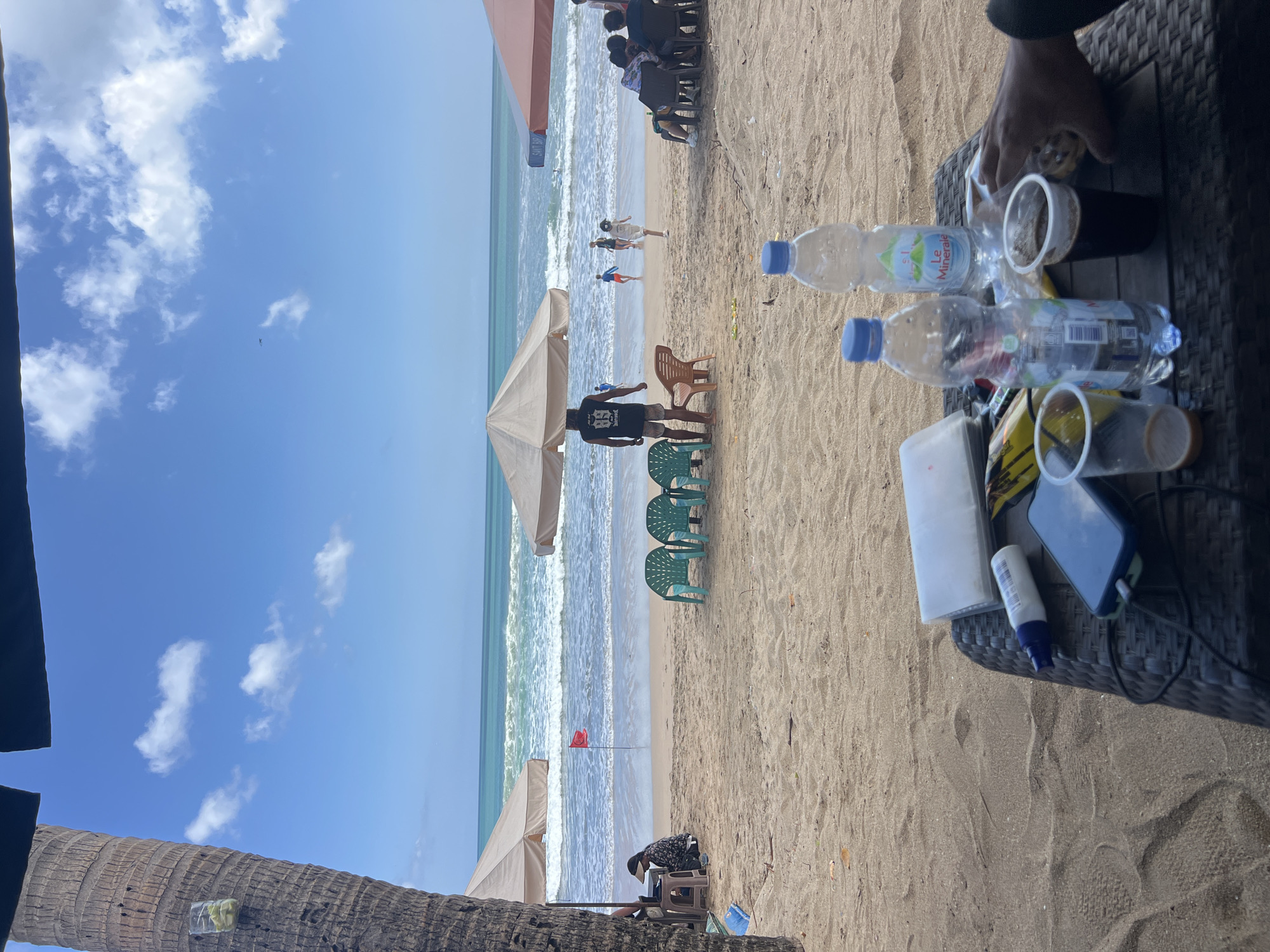 This is a medium-distance, eye-level shot of Kuta Beach, Indonesia, on a bright, partly cloudy day. In the foreground, a dark woven table holds various items, including two plastic cups with dark liquid, two plastic bottles of water, a smartphone, a tube of sunscreen, and a white rectangular object, possibly a packet of tissues. A person's right hand is visible on the right side of the frame, reaching towards the table. The table is set on the sandy beach, with the ocean and sky filling the background.
The middle ground of the image shows a sandy beach with several beach chairs and umbrellas. Two people are seated under a tan umbrella to the left, and a person in a black shirt with "88" on the back stands near a brown plastic chair in the center. To the right, a group of people are seated on chairs under a pink umbrella. Further back, on the wet sand near the ocean, several people are walking or playing in the water. A small red flag is planted in the sand near the water's edge, possibly indicating a warning. The turquoise ocean has moderate waves, and the sky is a bright blue with scattered white clouds. A palm tree trunk is visible on the far left of the frame, partially obscuring the scene.