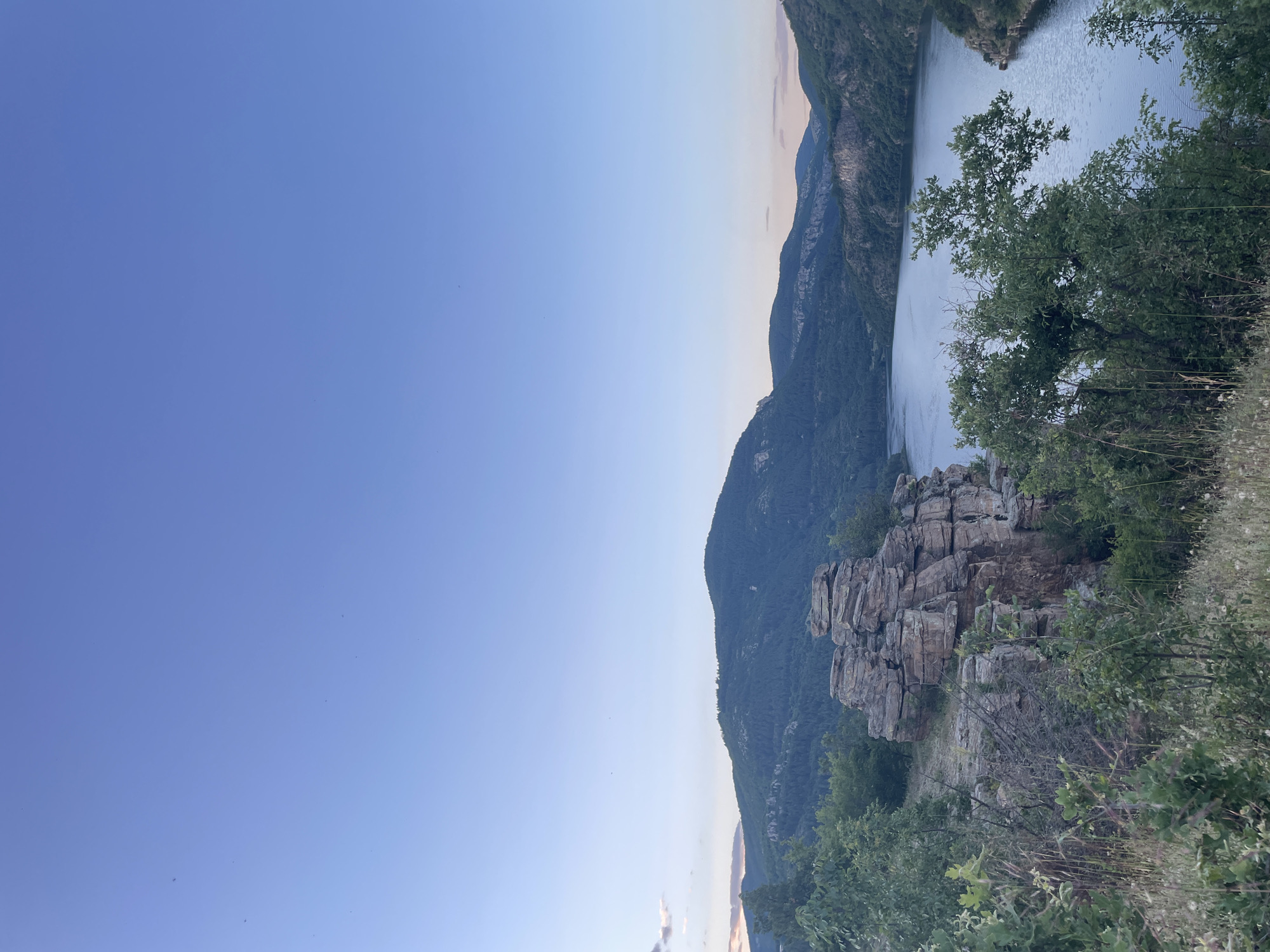 This is a vertical, eye-level, outdoor, wide shot of a serene natural landscape at dusk, likely in the vicinity of Suhovo, Bulgaria. The image features a tranquil body of water, resembling a reservoir or a lake, winding through a valley. The water is calm, reflecting the soft hues of the twilight sky.
Dominating the foreground to the left is a prominent rock formation, appearing as a cluster of weathered, layered stones. Lush green vegetation, including trees and grasses, surrounds the rocks and slopes down towards the water. In the background, rolling hills and mountains are covered in dense forests, their outlines softened by the fading light. The sky is a clear, gradient blue, transitioning to lighter tones near the horizon where a few wisps of clouds catch the last light of the setting sun. There are no people or animals visible in the scene. The overall atmosphere is peaceful and natural.