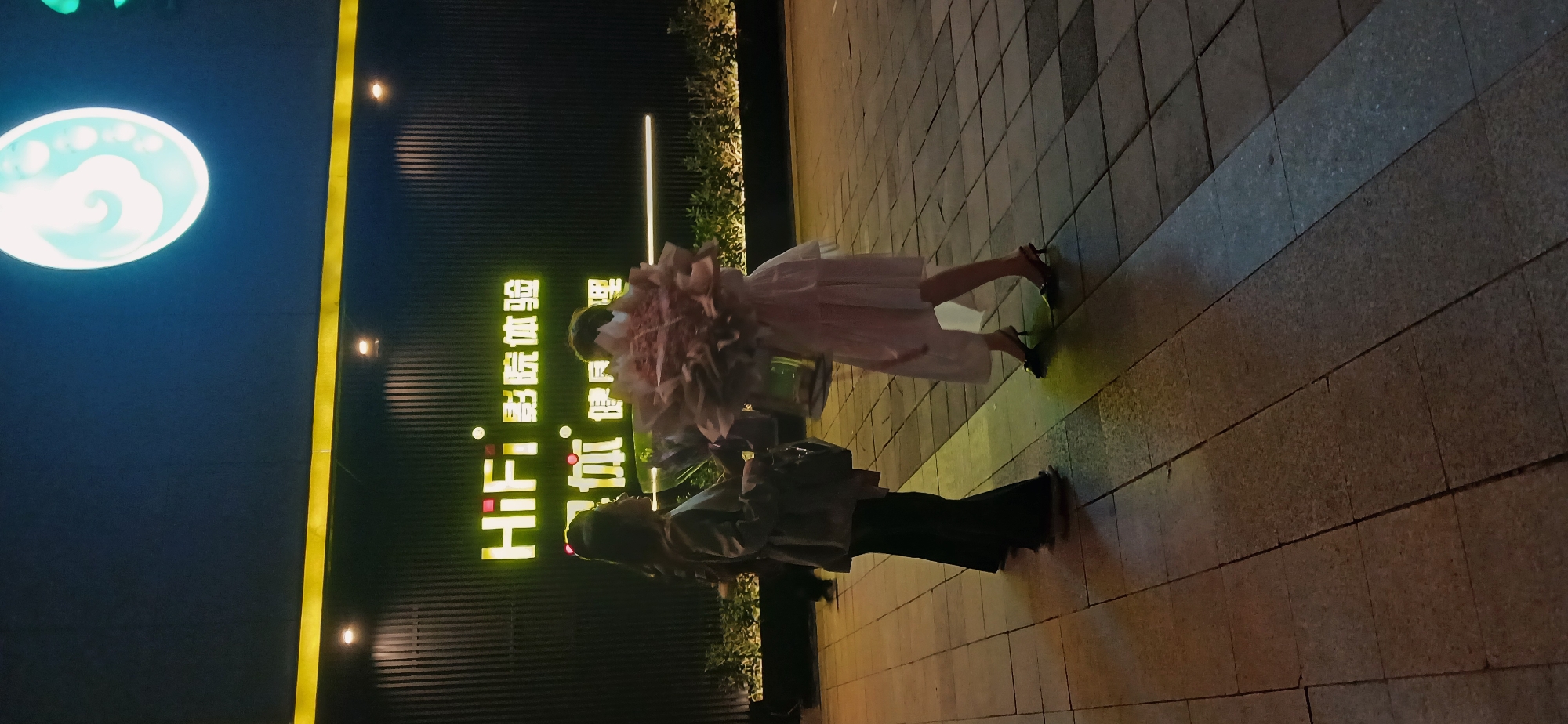 Two women are standing on a paved walkway. One woman is holding a large bouquet of flowers and is wearing a white dress and black heels. The other woman is wearing black pants and a light-colored jacket and is holding a bag. Behind them is a building with a sign that reads "HIFI. 渝洲新零售体验店" in Chinese characters. A yellow stripe runs vertically along the side of the building. To the left of the building is a circular sign with a cloud design. The image was taken at night in Chongqing High-tech Zone, China.