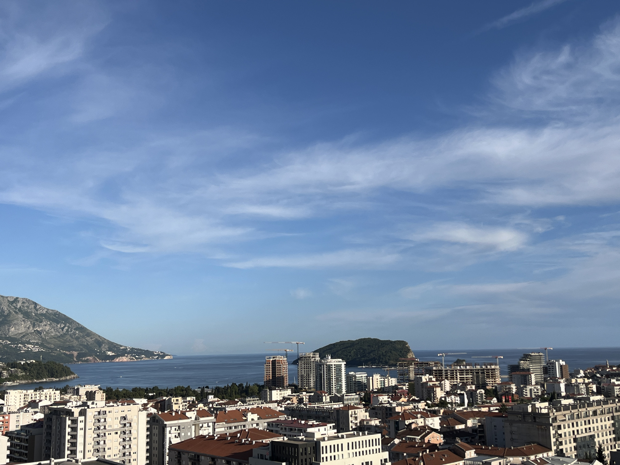 The image shows a cityscape of Budva, Montenegro, taken from a high vantage point. The foreground is dominated by a dense collection of buildings, predominantly white or light-colored, with red-tiled roofs on many structures. In the midground, the Adriatic Sea is visible, with an island in the distance covered in green vegetation. Several construction cranes are present amongst the buildings, indicating ongoing development. To the left, a mountain range is partially visible, sloping down towards the sea. The sky is a gradient of blue, with wispy clouds scattered throughout. The overall scene suggests a sunny day in a coastal urban environment.