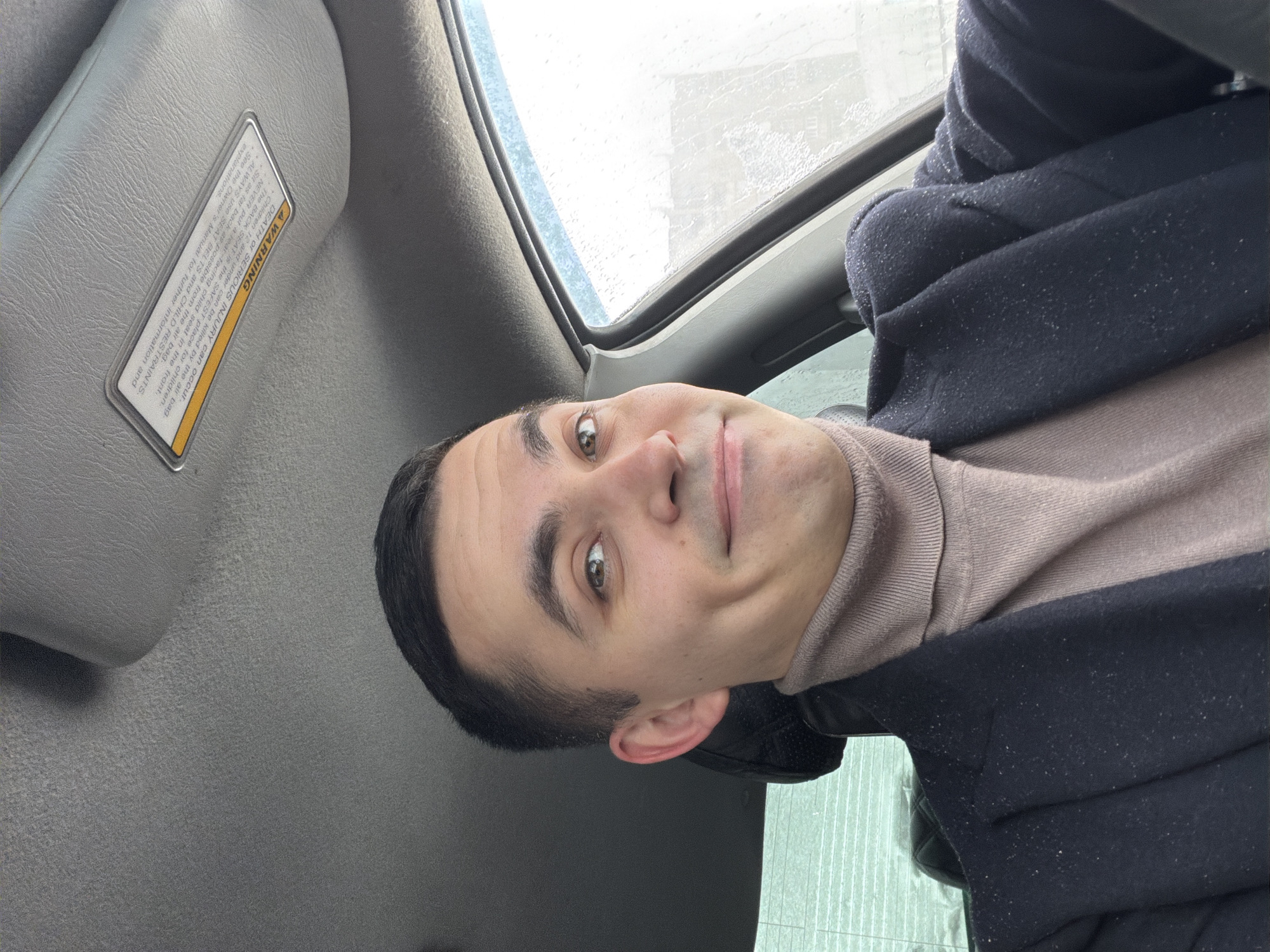 A man is sitting in a car, seemingly in the driver's seat, as indicated by the sun visor labeled with a warning about airbags located to his left. The view through the car window is obscured by what appears to be rain or condensation. He's wearing a turtleneck sweater and a dark coat. The image provides no location information about Izhevsk, Russia.