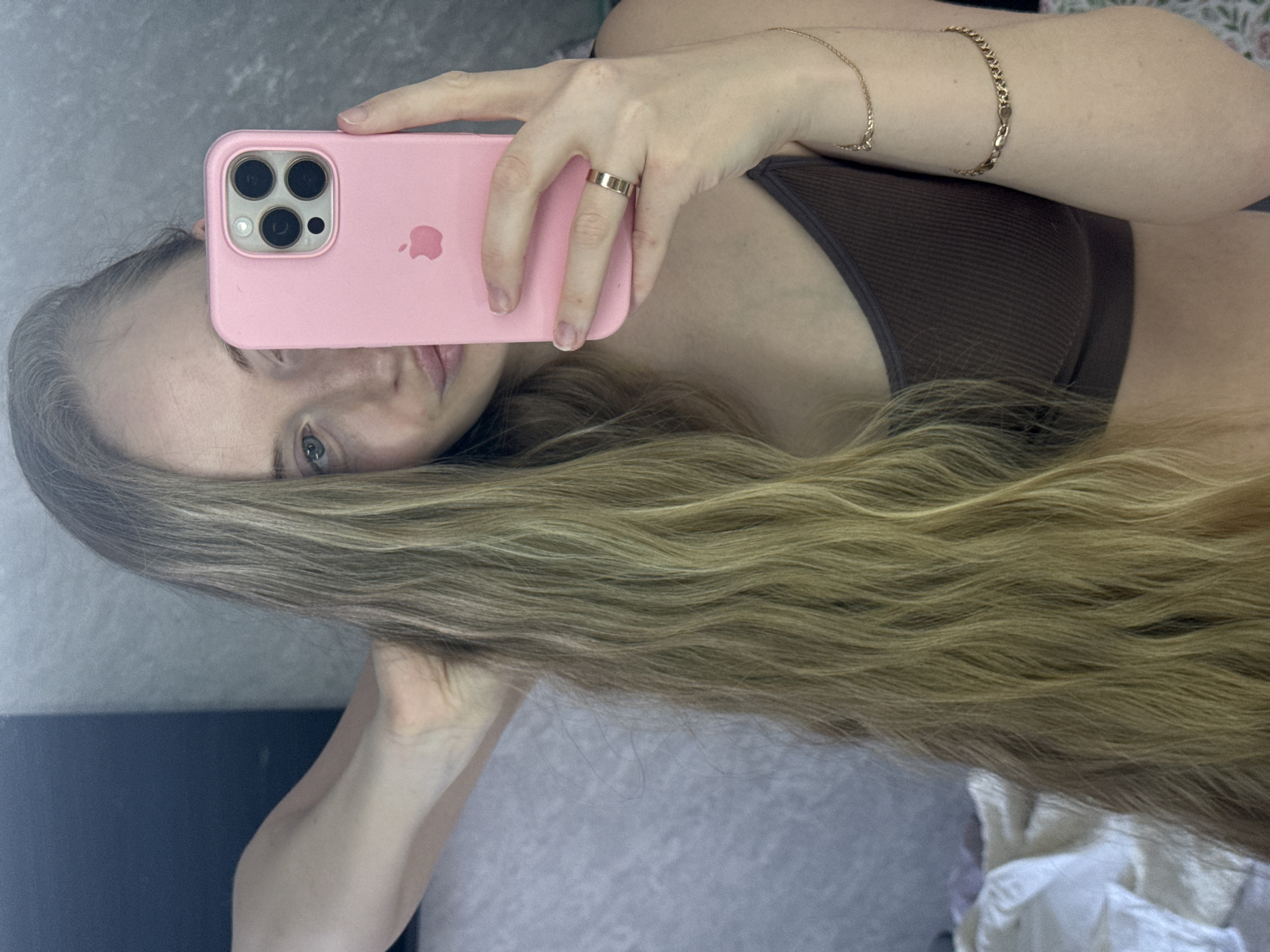 A woman is holding a pink iPhone in her right hand, taking a selfie. She is wearing a brown top. She also has a gold ring on her finger and two gold bracelets on her right wrist. Her blonde hair is draped over her arm. The background is a gray wall. There is no indication of the location being in the city of Petropavlovsk-Kamchatsky, Russia.
