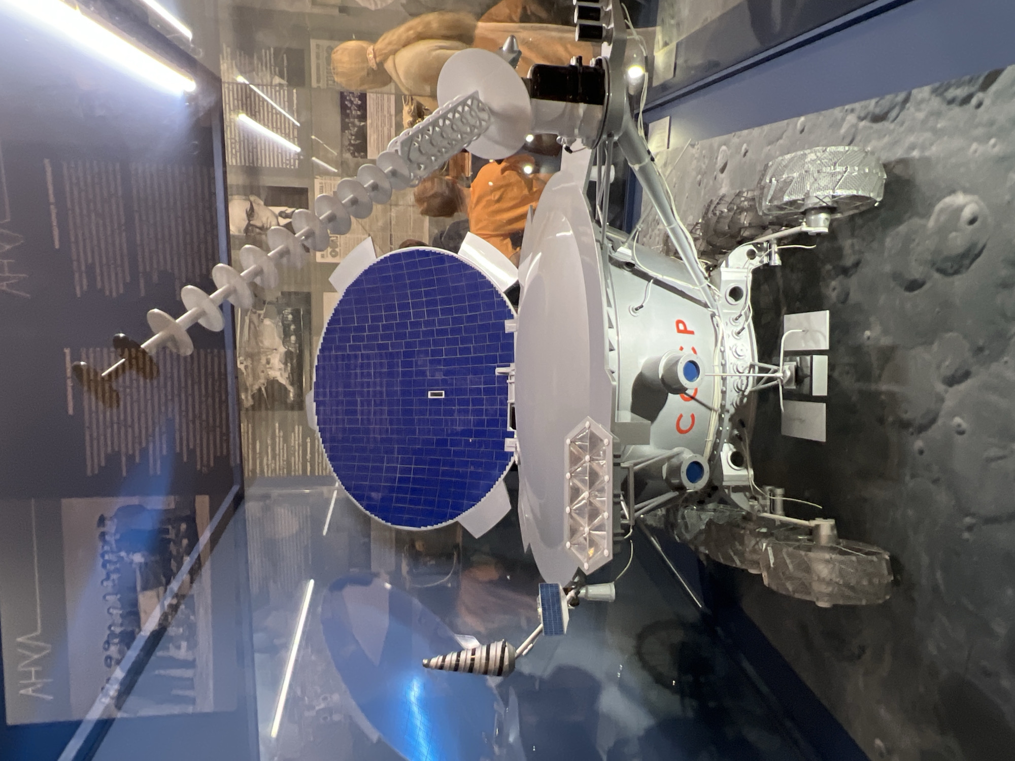 The image shows a model of the Soviet lunar rover Lunokhod on display at the Museum of Cosmonautics and Rocket Technology in Saint Petersburg, Russia. The model features a large blue solar panel, a dish antenna, and several other scientific instruments. The rover is shown against a backdrop of a lunar surface. The display case is made of glass.
